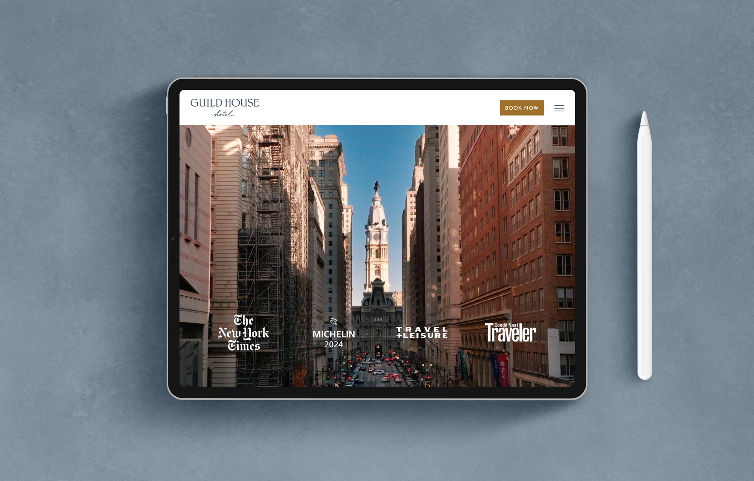 GHH-Tablet A tablet displaying the Guild House Hotel website featuring a city street view of Philadelphia with a historic buildings and skyscrapers in the distance. Logos of The New York Times, Michelin 2024, Travel + Leisure, and Traveller are visible. A stylus is placed beside the tablet.