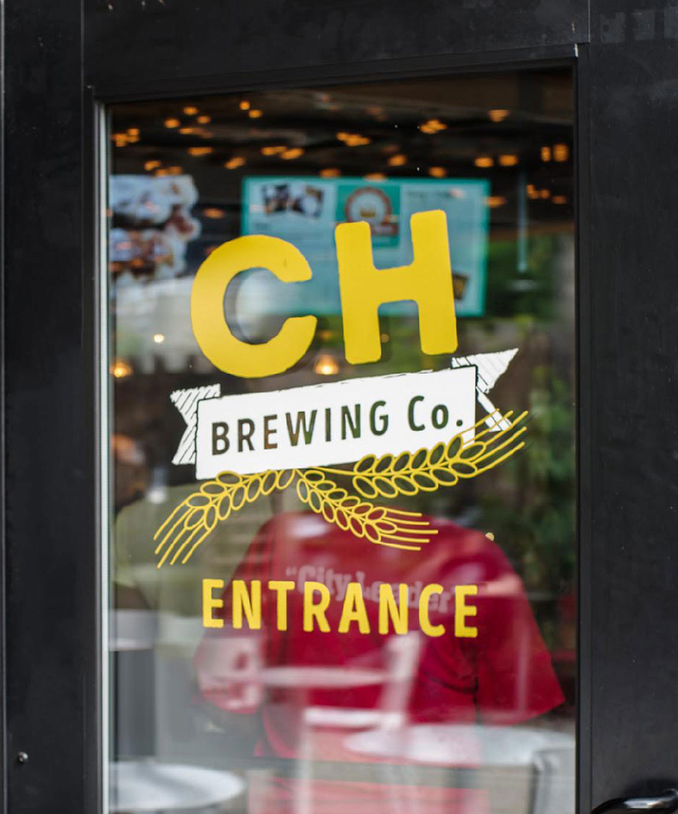 CHBC-Entrance Glass door with "CH Brewing Co. Entrance" in bold yellow letters and a wheat illustration, reflecting a cozy, inviting interior with soft lighting.
