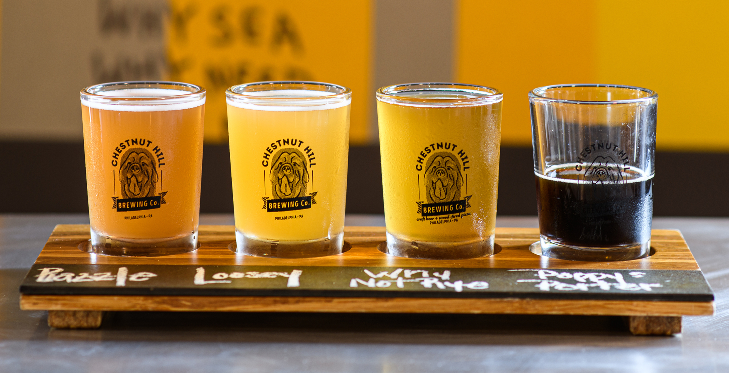CHBC-Flight Four beer glasses of varying colors sit on a wooden flight board, labeled "Razzle," "Loosey," "Why Not Rye," and "Poppy Porter" from Chestnut Hill Brewing Co.