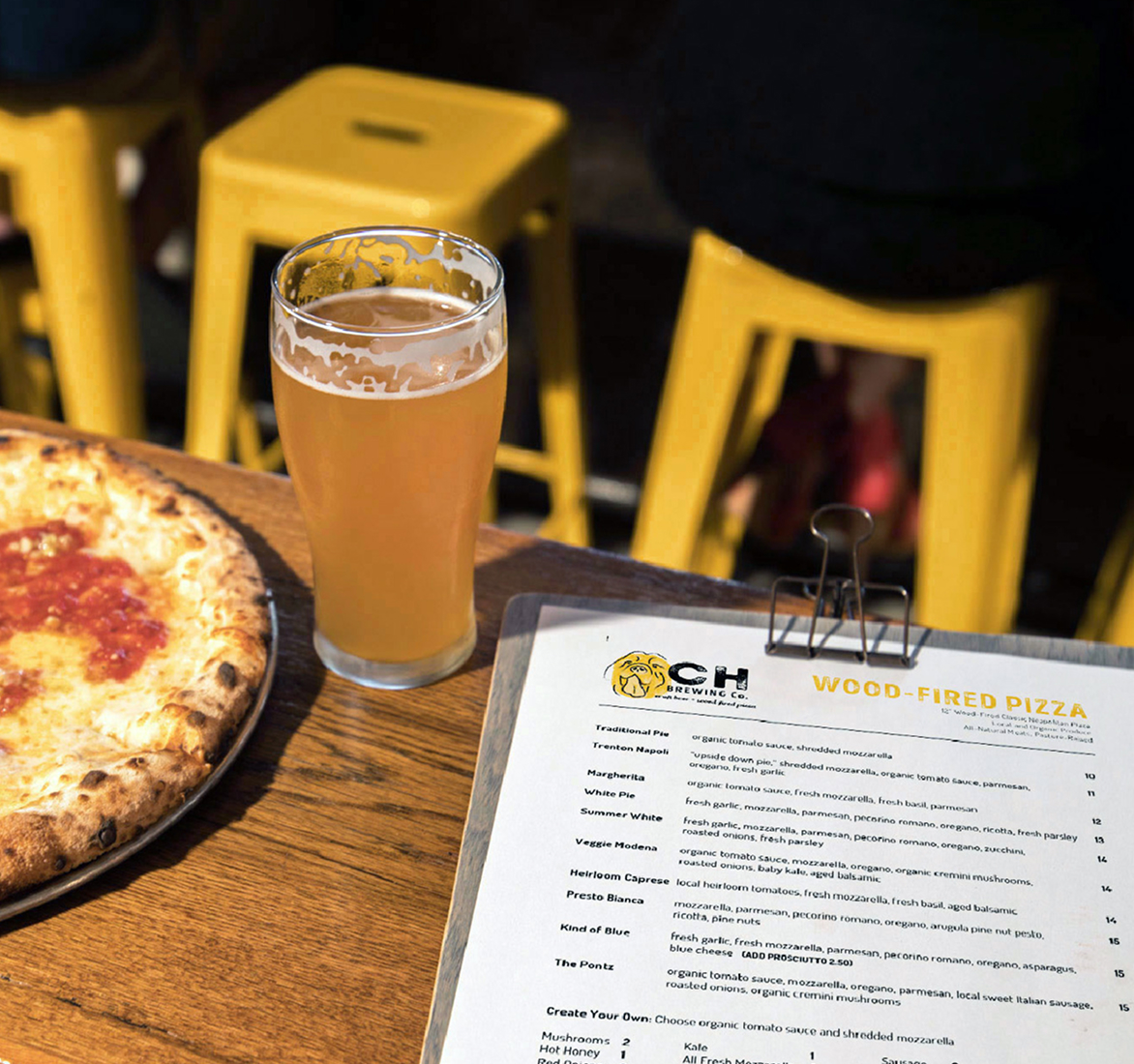 CHBC-Menu A wooden table with a menu labeled "Wood-Fired Pizza," a glass of pale beer, and a partially visible pizza. Yellow stools are in the background, creating a casual dining atmosphere.