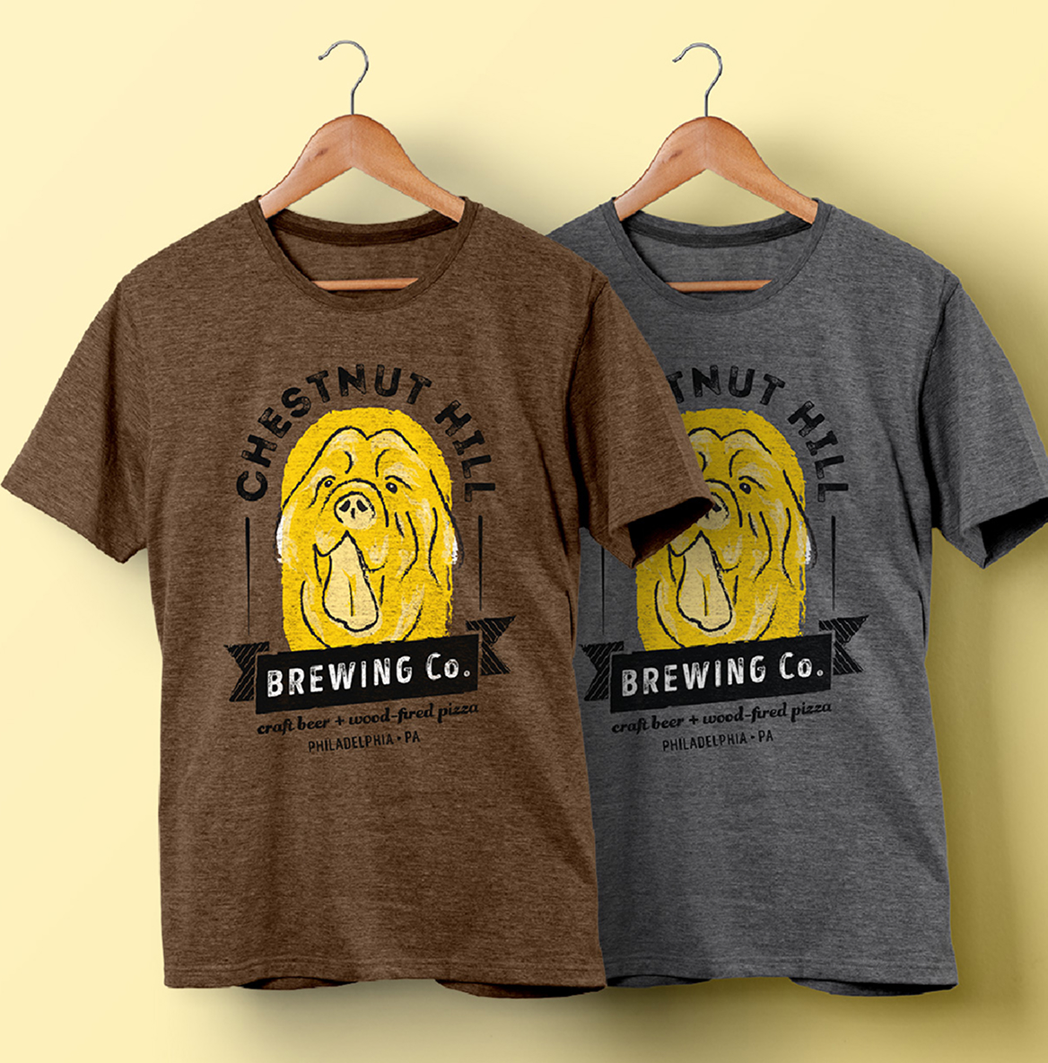 CHBC-Shirts Two t-shirts hang side by side, one brown and one gray, both featuring a yellow dog illustration and the text "Chestnut Hill Brewing Co."