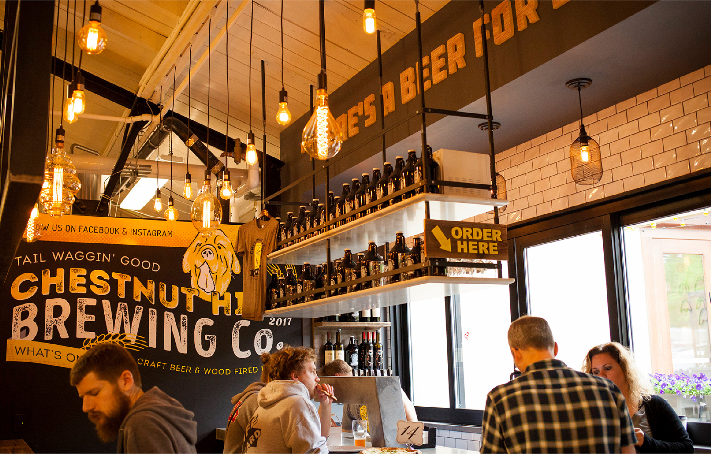 CHBC-Store A cozy brewery interior with warm lighting, patrons at a counter, and rows of beer bottles. A sign reads "Chestnut Hill Brewing Co." and "Order Here."