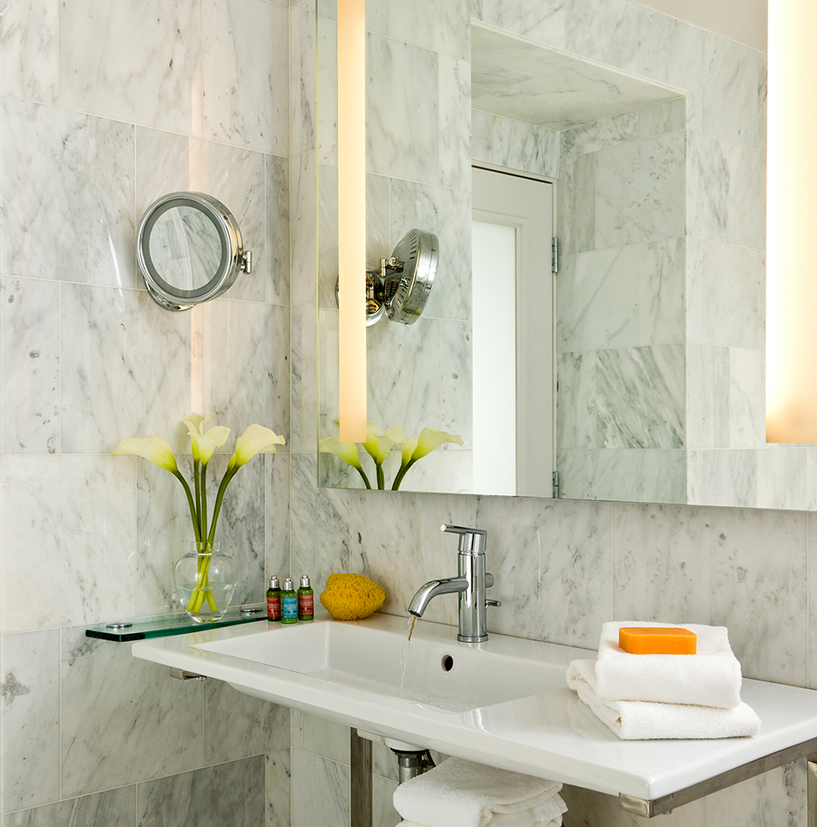 Elegant bathroom with marble walls and a modern sink. A mirror reflects calla lilies in a vase. Soft lighting and folded towels create a serene ambiance.