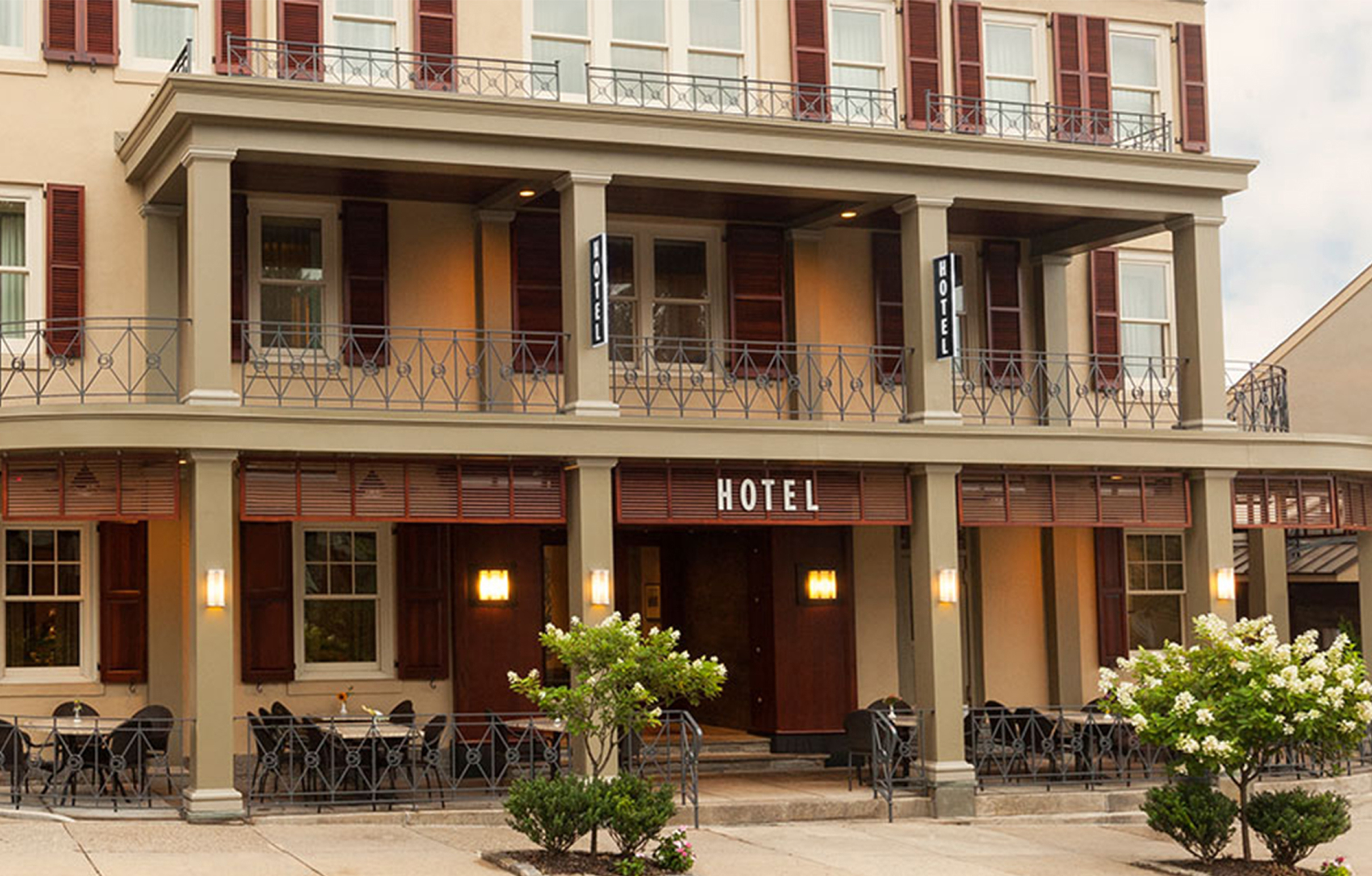 Hotel facade with a large balcony, labeled "Hotel" above the entrance. Outdoor seating, flowers, and warm lighting create an inviting atmosphere.