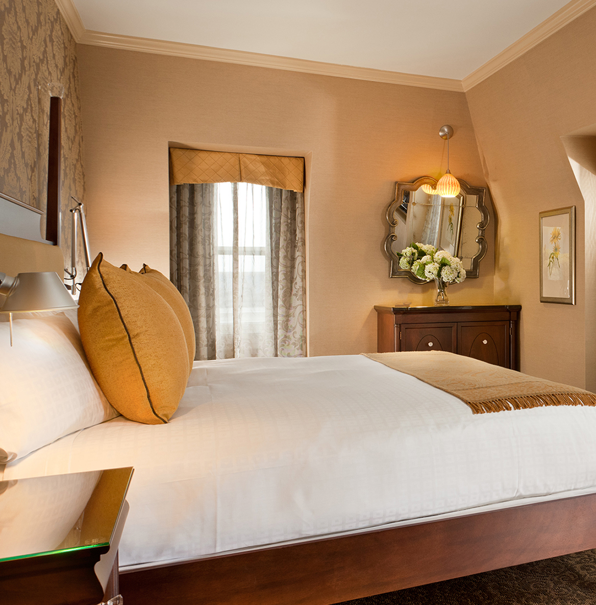Elegant hotel room with a cozy bed, brown pillows, and a warm beige theme. Features ornate mirror, soft lighting, and floral decor, creating a serene ambiance.