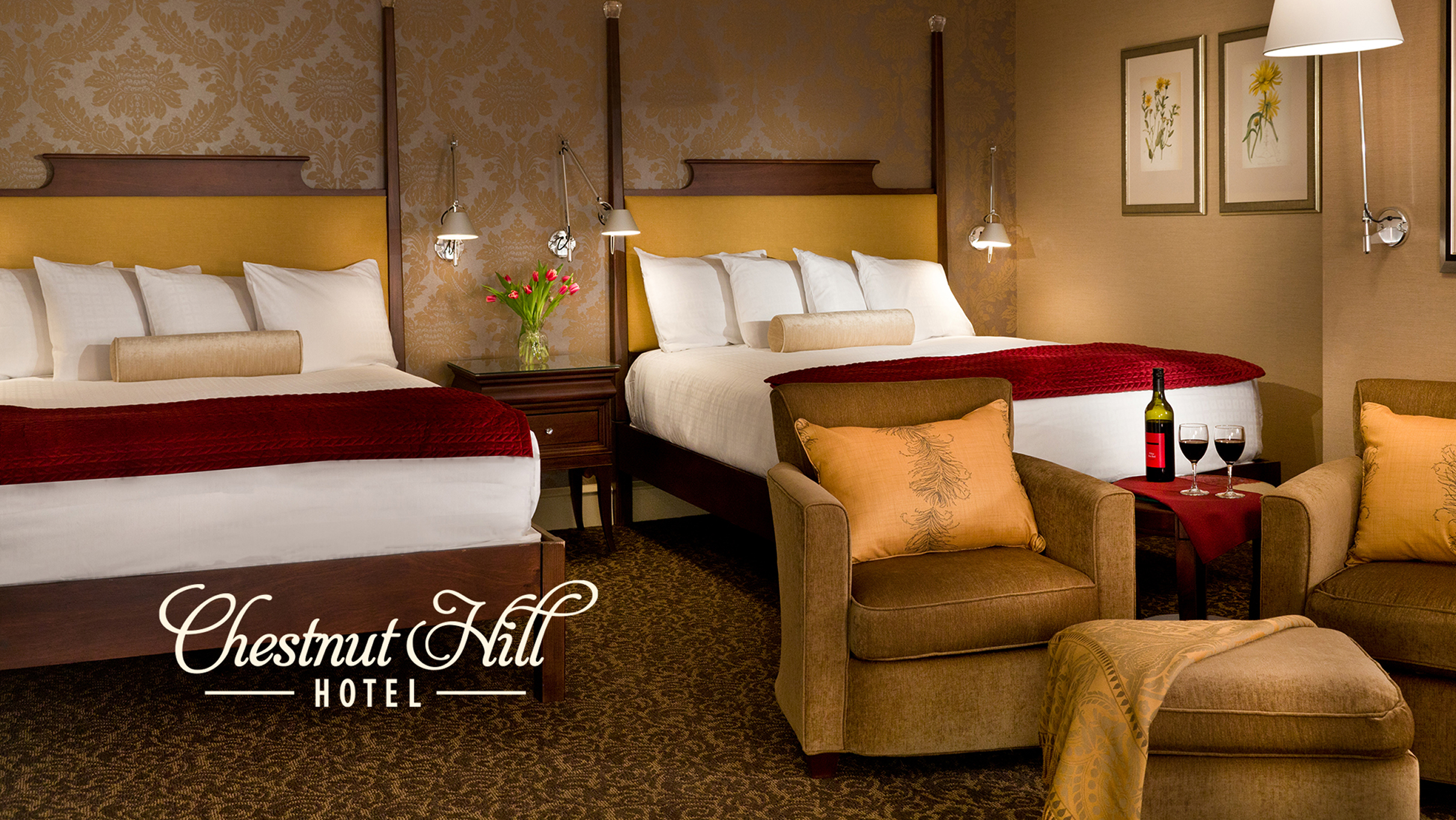 Elegant hotel room with two double beds, rich red and gold accents, and plush armchairs. A cozy atmosphere with wine and flowers enhances its warmth.