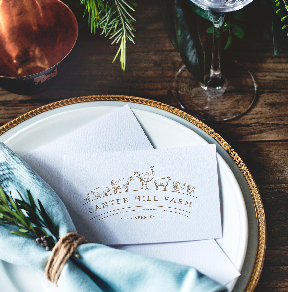 Canter Hill Farm-Plate Elegant table setting with a white card reading “Canter Hill Farm.” A blue napkin with rosemary is on a white plate with a gold rim, surrounded by greenery.