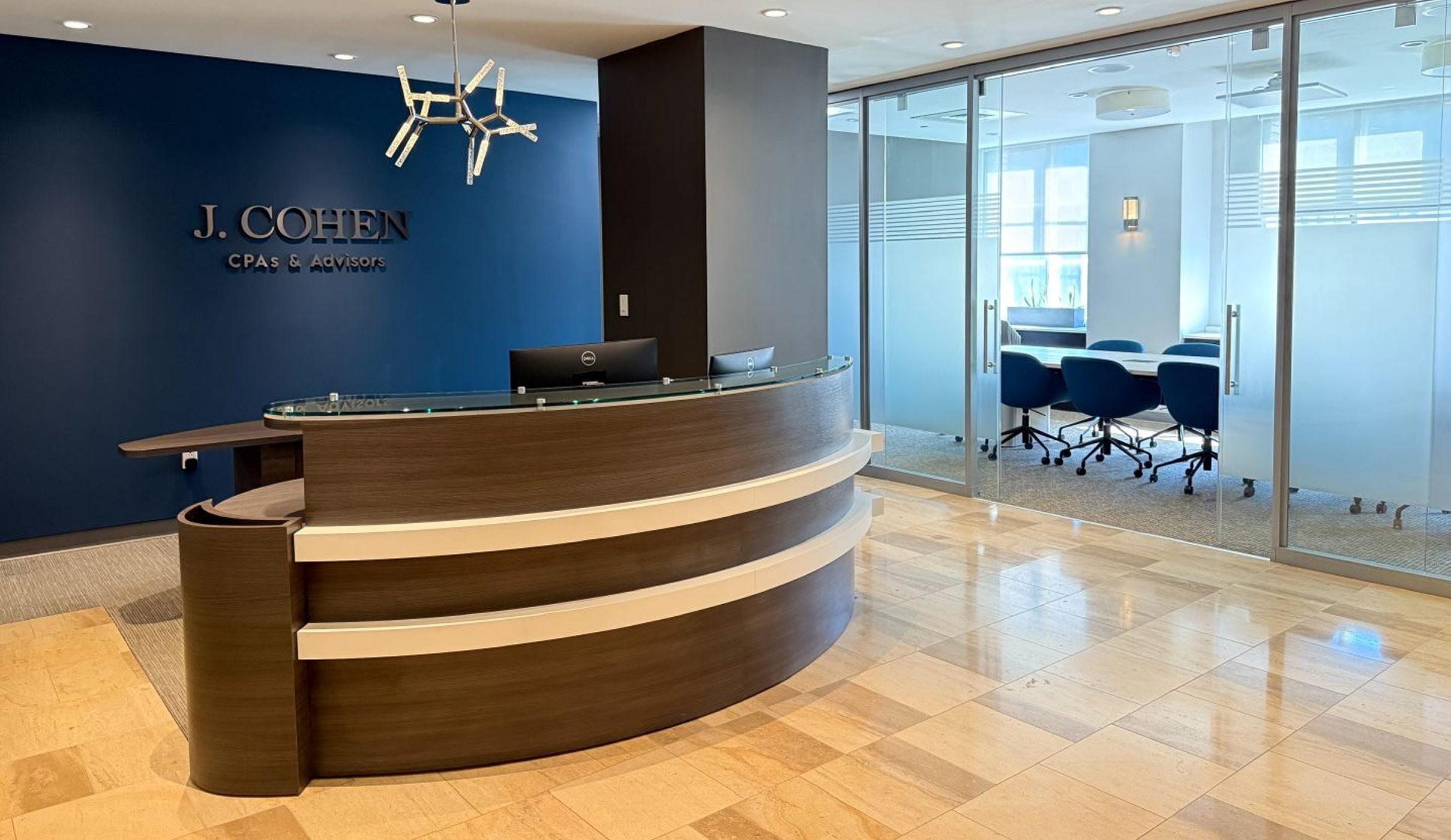 Modern office reception with a sleek curved desk, dark blue accent wall, and stylish light fixture. The wall has the "J. Cohen CPAs & Advisors" logo. Glass doors reveal a conference room with blue chairs.