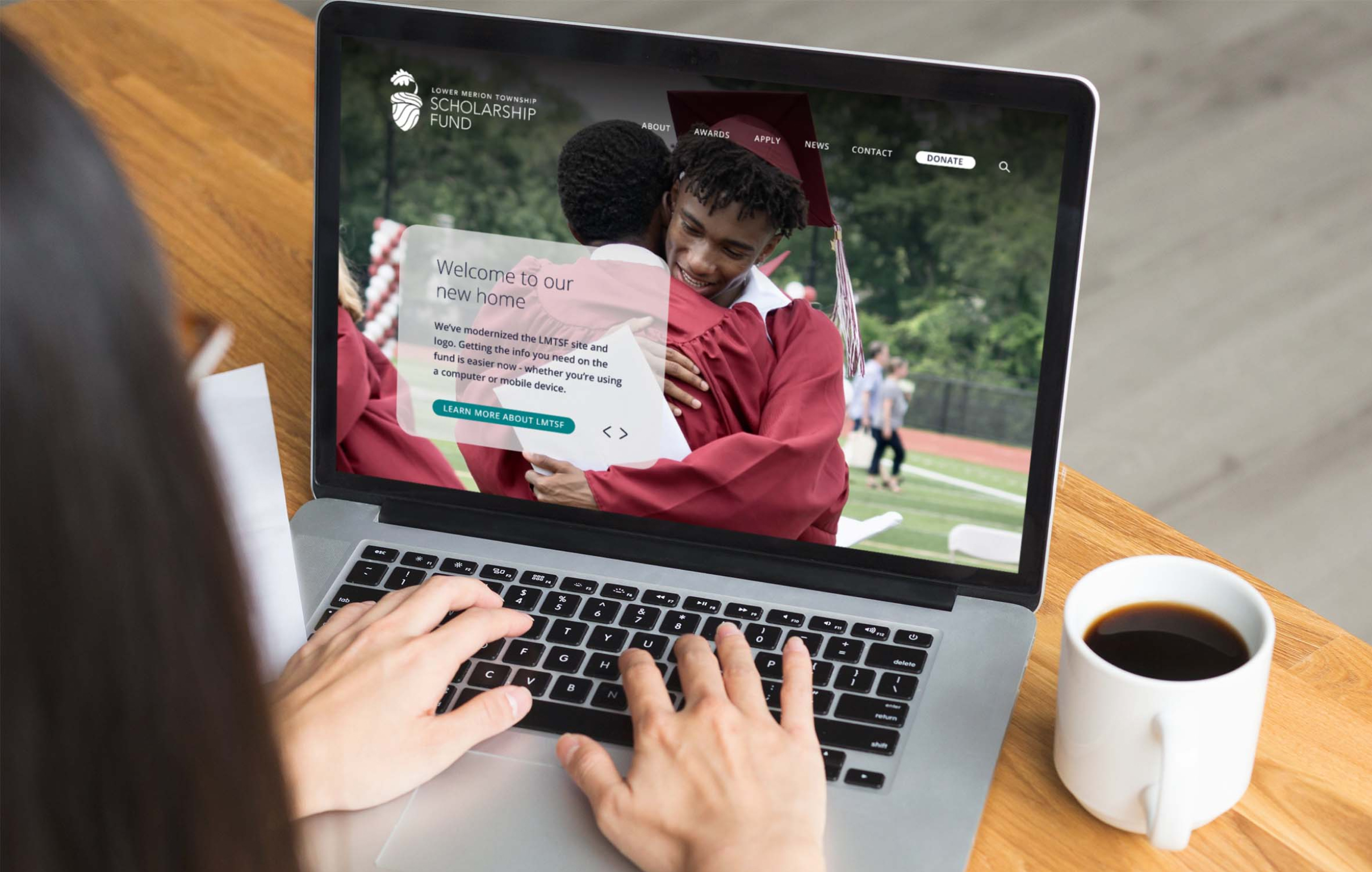 A person views the LMTSF website on a laptop showing two graduates hugging in red gowns and caps. The scene conveys celebration and achievement. A coffee cup sits nearby.