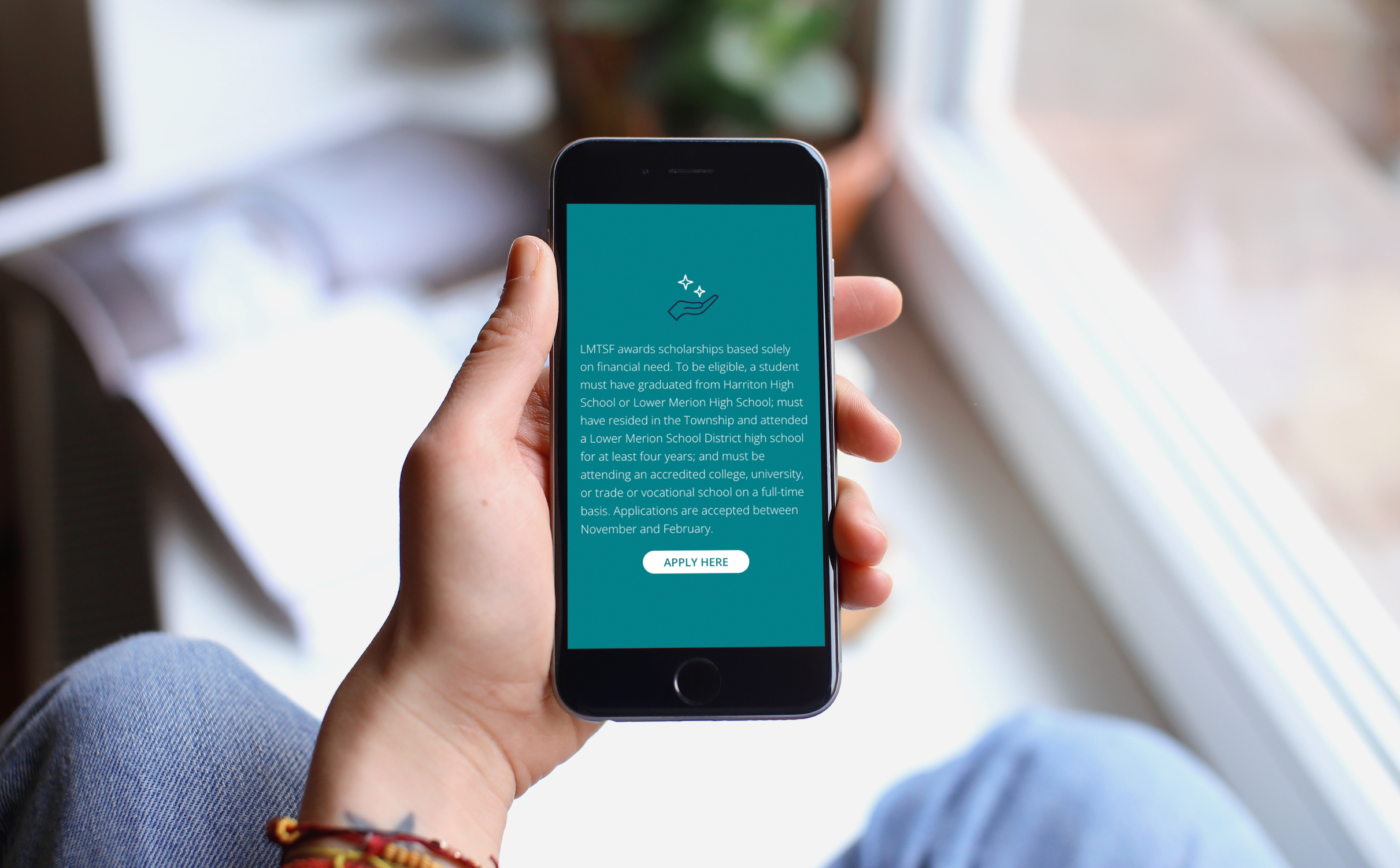 A person holds a smartphone displaying a scholarship application screen. The background is a cozy room by a bright window, creating a calm and focused atmosphere.