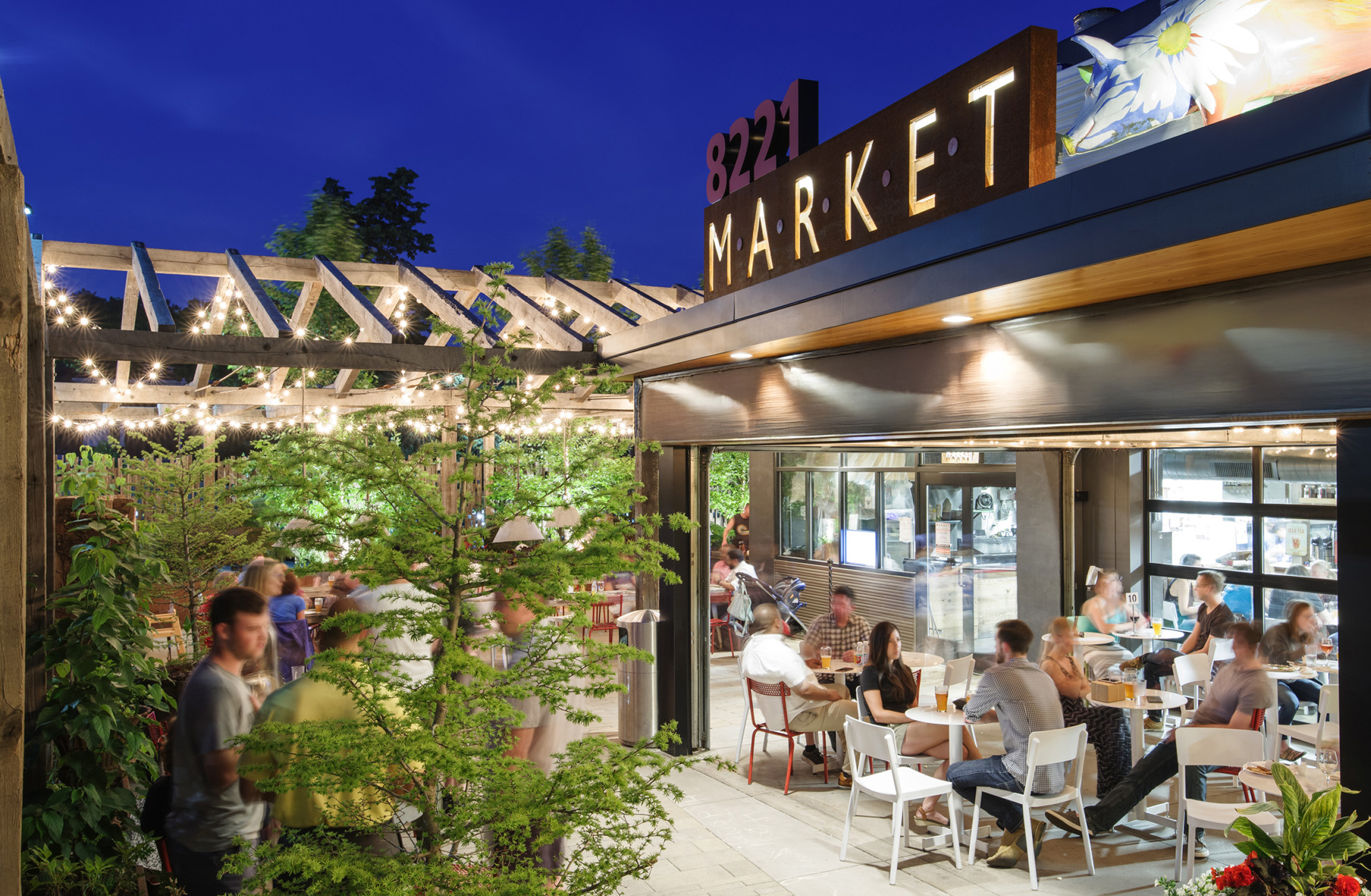 A lively market scene at dusk, with people dining outdoors under string lights and a wooden trellis. The sign reads "8221 MARKET," creating a warm, inviting atmosphere.