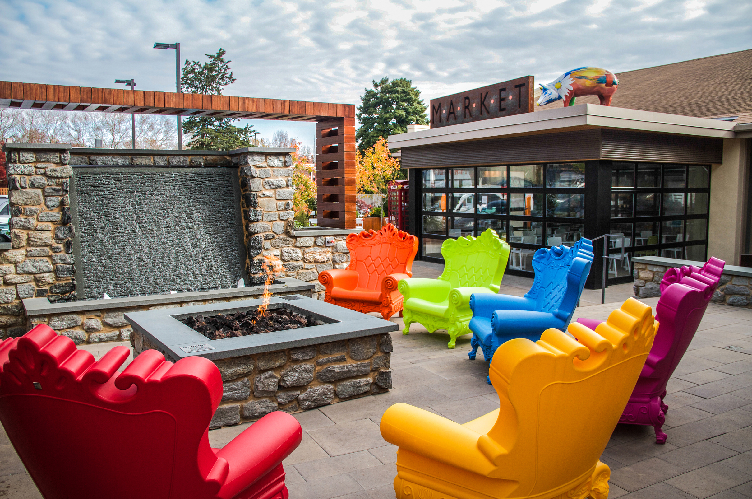Vibrant outdoor seating features colorful chairs around a stone fire pit near a waterfall wall. A "Market" sign is in the background, evoking a lively atmosphere.