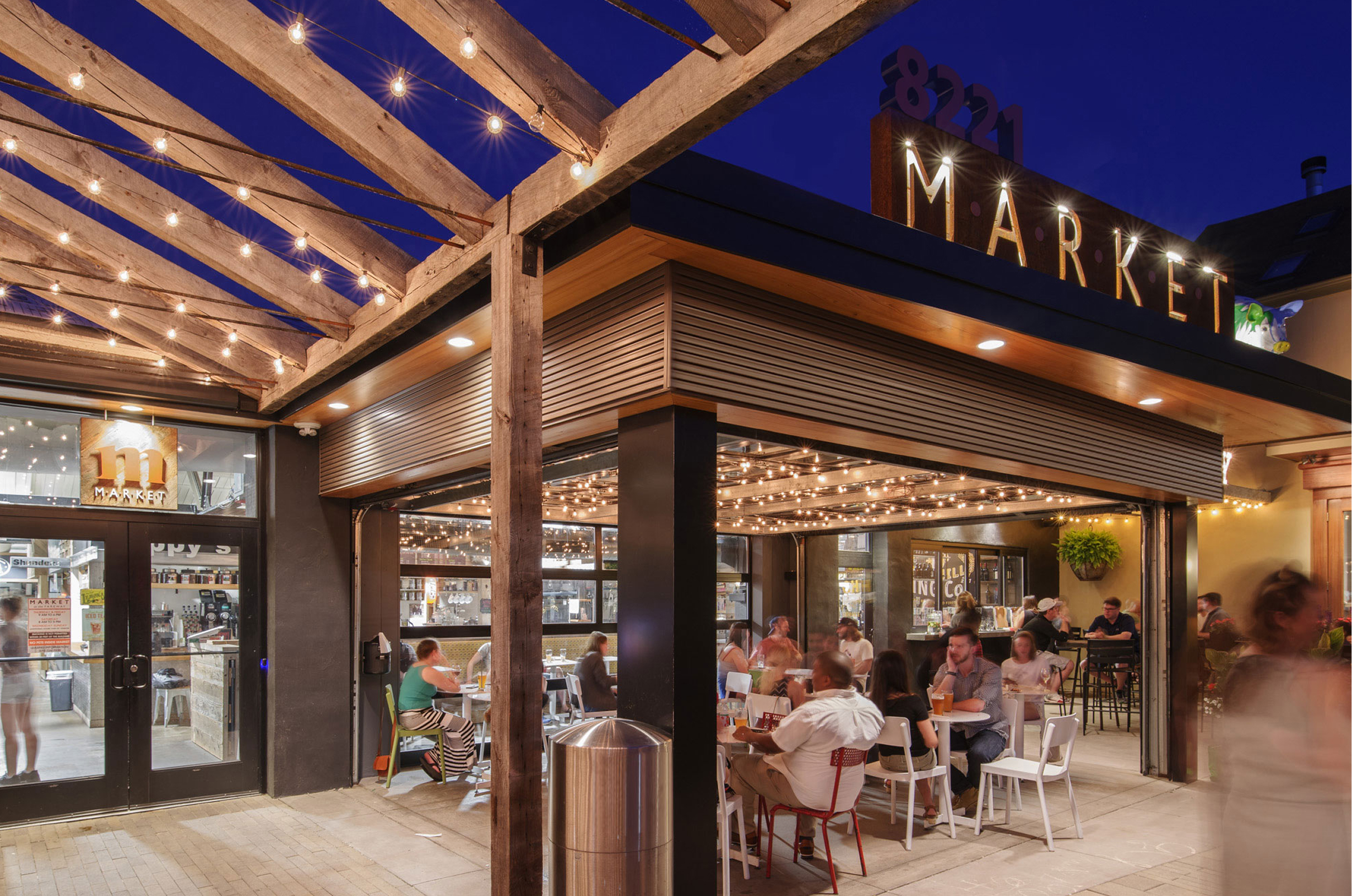 Bustling outdoor market dining area at night, warmly lit by string lights. People sit and converse at tables under a wooden pergola, creating a lively atmosphere.