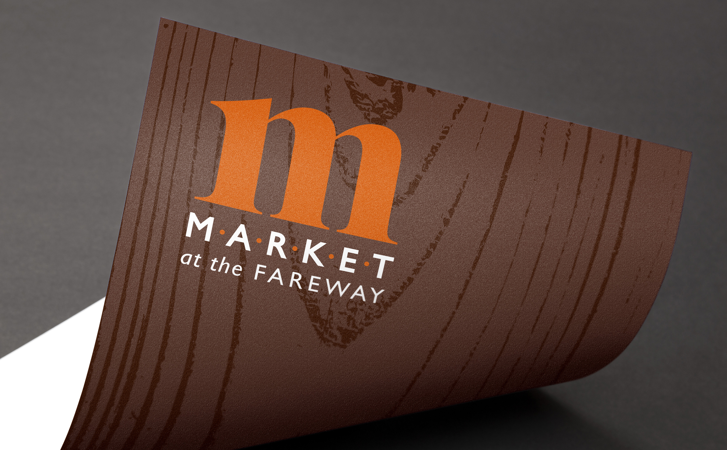 Logo reading 'Market at the Fareway' in white text on a brown wood-textured background. An orange lowercase 'm' is prominently featured above the text.