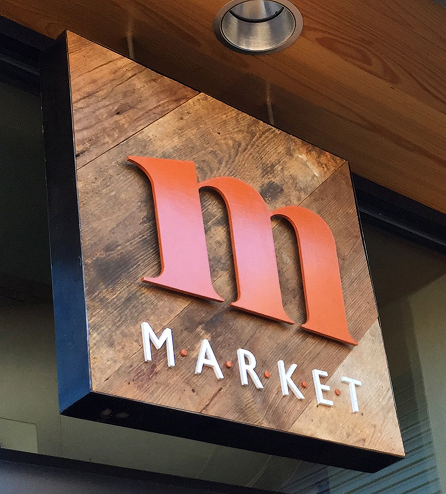 Sign reads "Market" with a large orange "m" on a wooden background. The tone is rustic and inviting, with warm wood and bright letters.