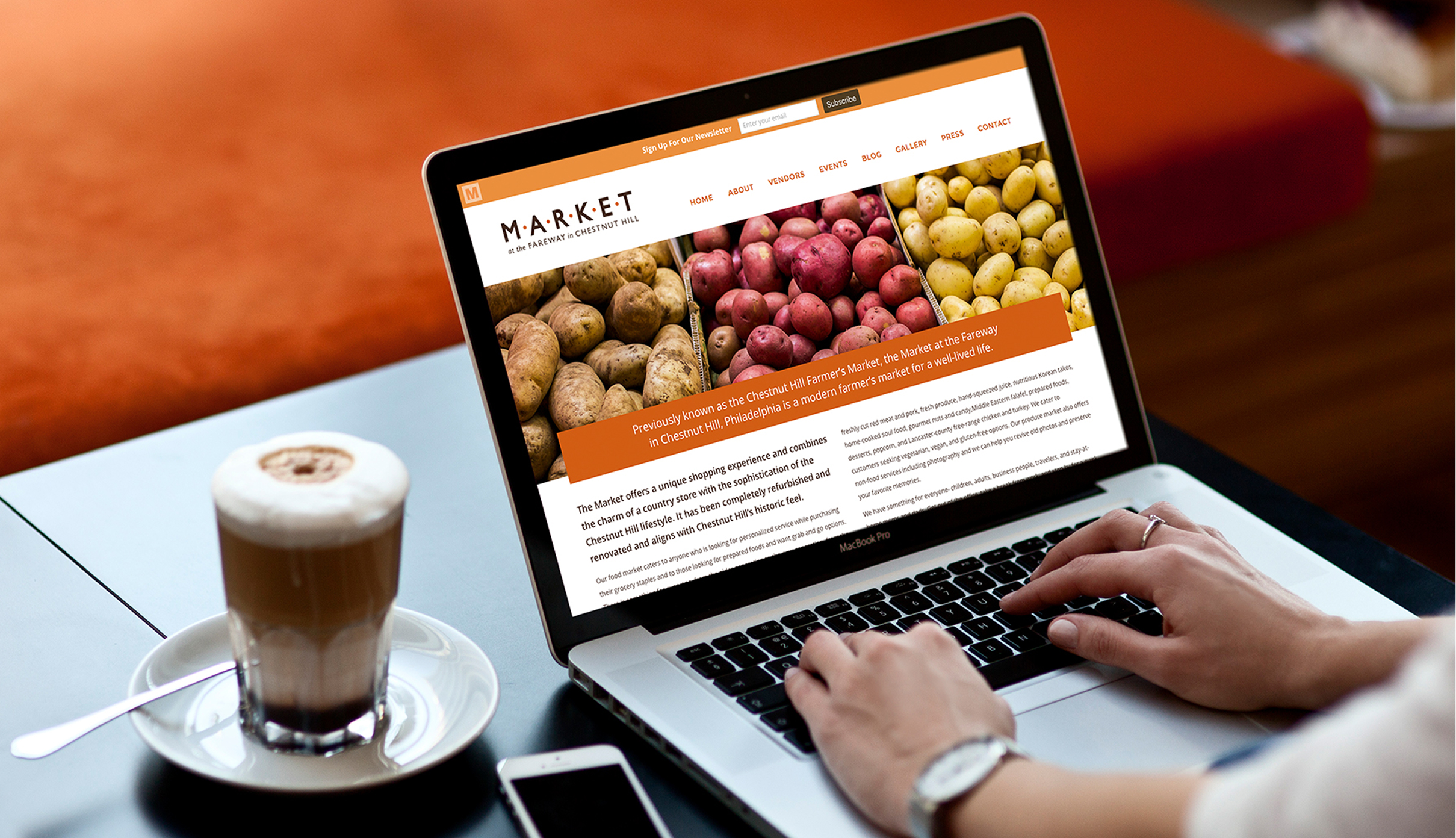 A person uses a laptop displaying the Market at the Fareway website featuring colorful potatoes. A latte sits nearby, creating a cozy, focused work atmosphere.
