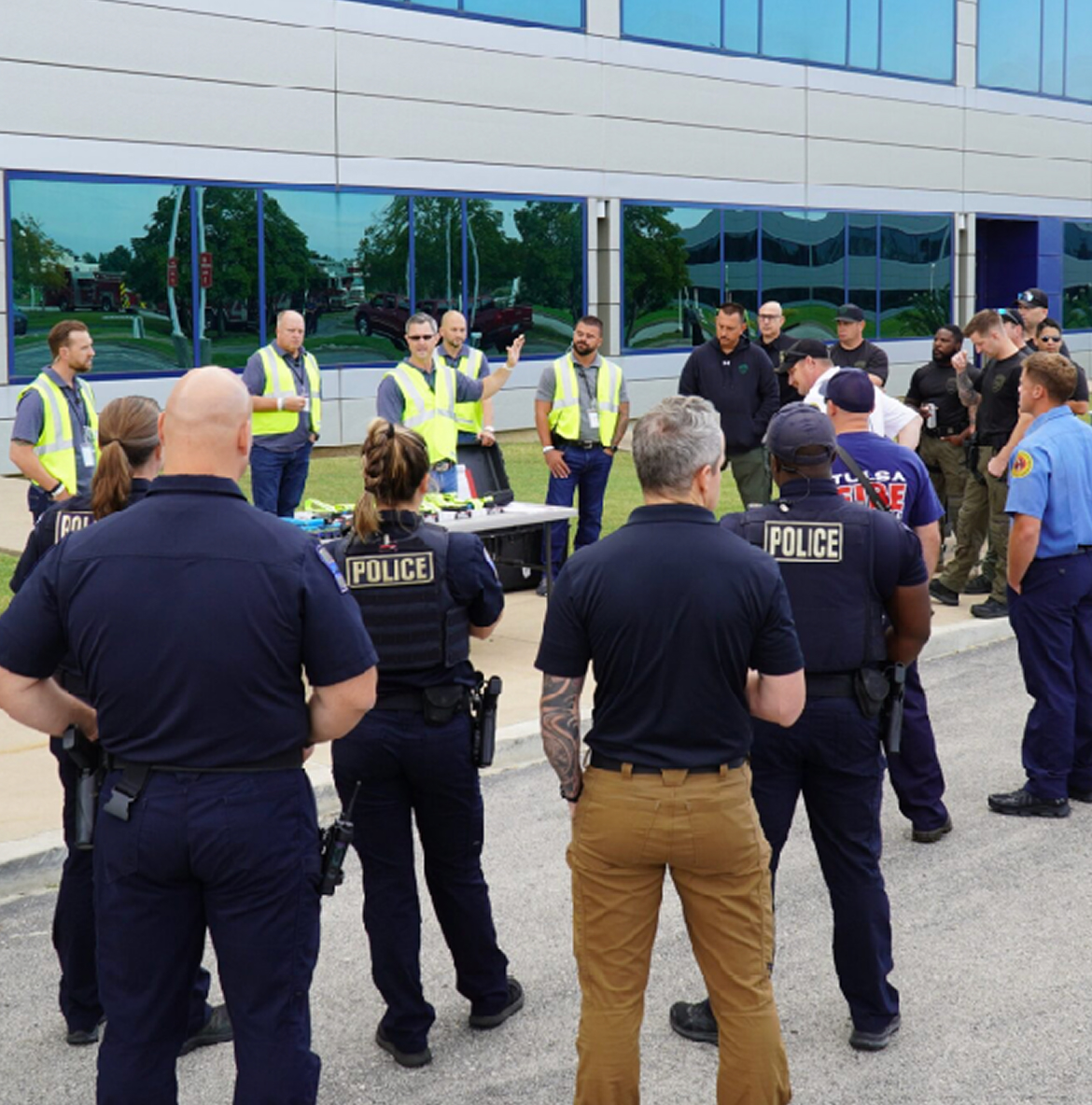 Tomahawk-Event A group of police and emergency personnel gather outdoors for training. Instructors in yellow vests stand by a table, gesturing as they present.