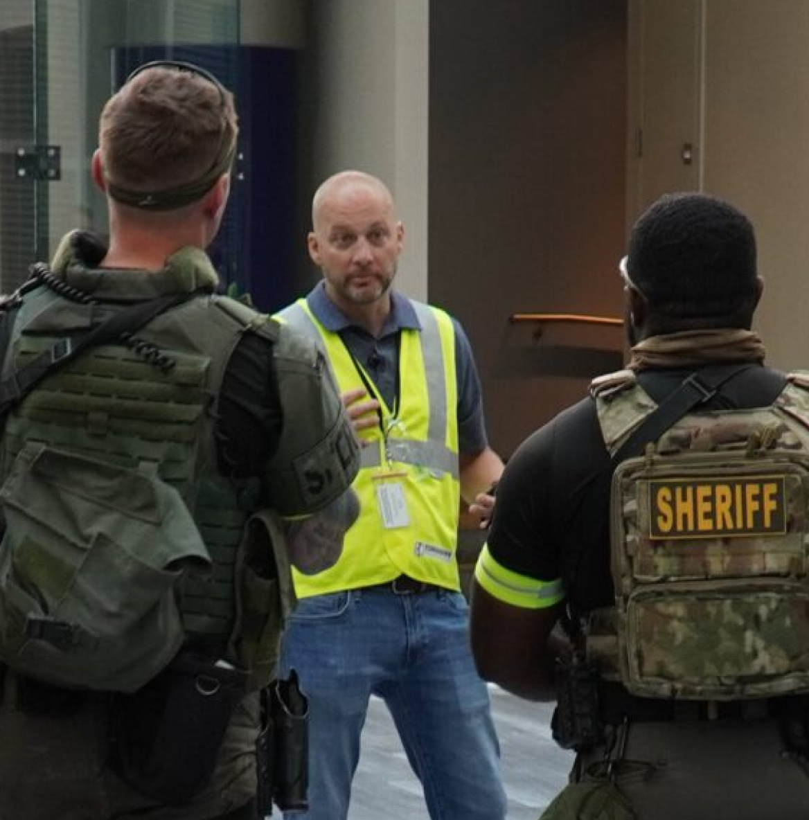 Tomahawk-Training A man in a yellow safety vest speaks to two sheriff officers in tactical gear. The tone is serious, suggesting a briefing or discussion.
