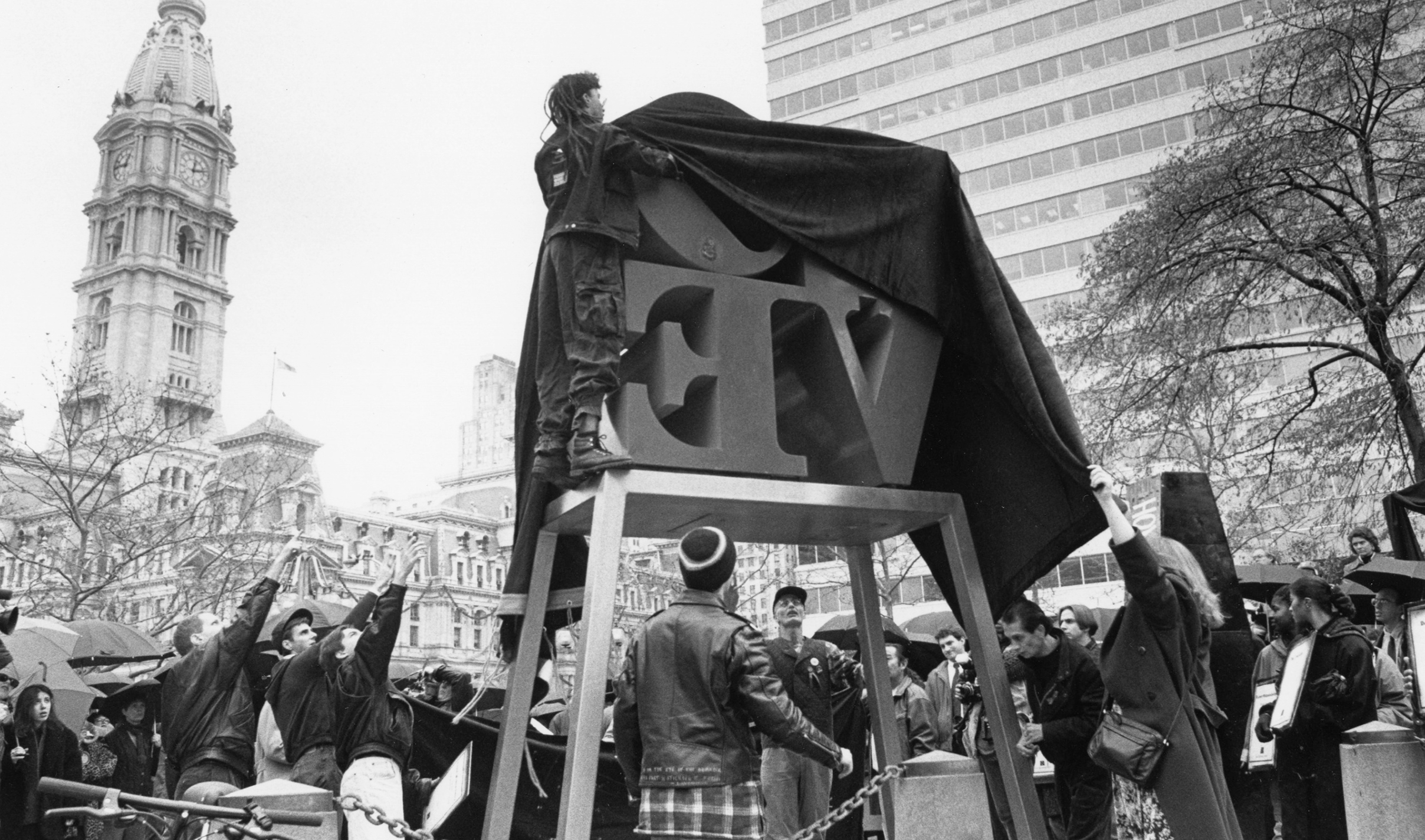 Crowd gathers as activists unveil the "LOVE" statue under a tarp in a Philadelphia, with historic architecture and a tall building in the background. The mood is energetic and anticipatory.
