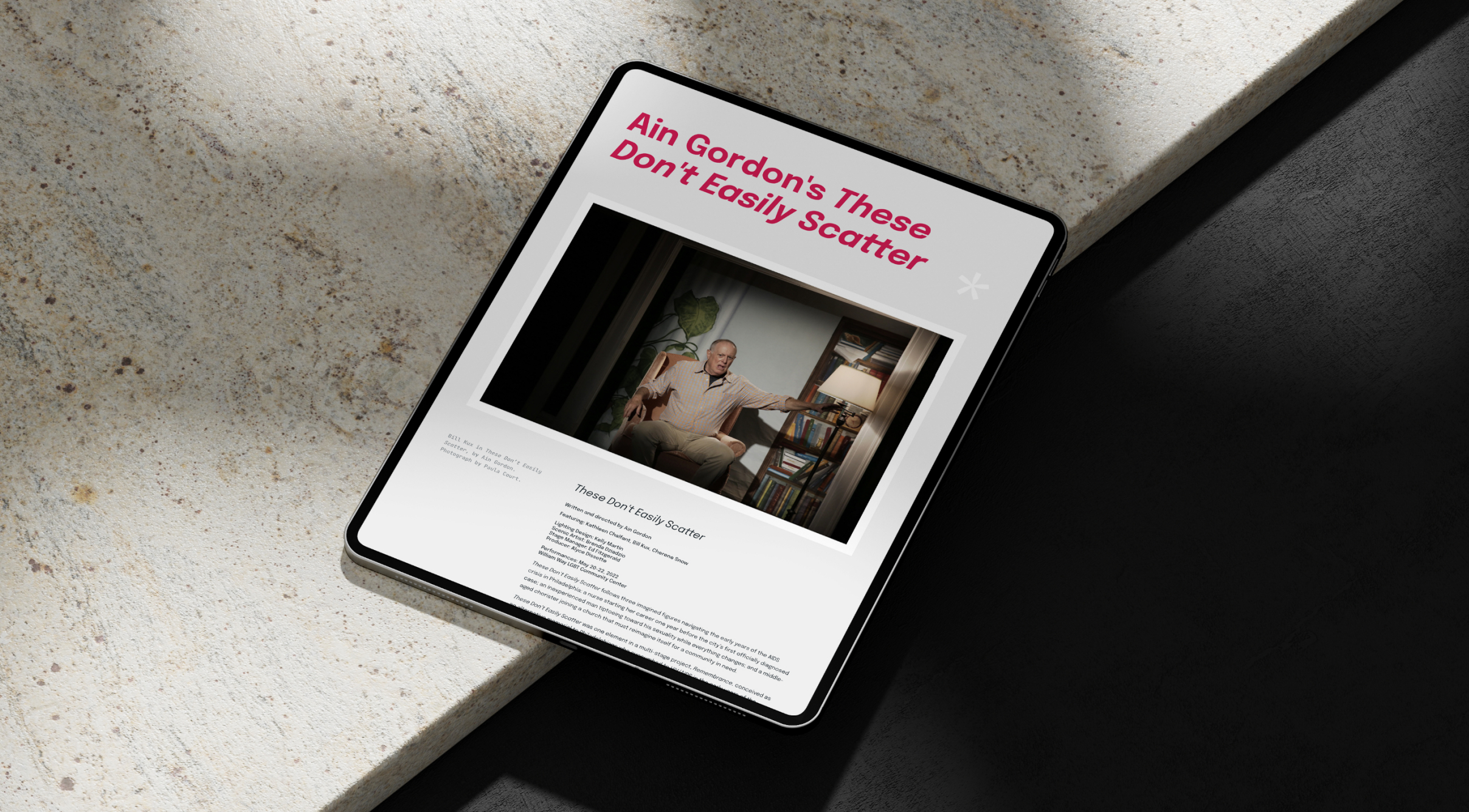 A tablet displaying a webpage for a performance titled "Ain Gordon's These Don't Easily Scatter" with a photo of a seated man gesturing. It's placed on a stone surface with soft lighting.