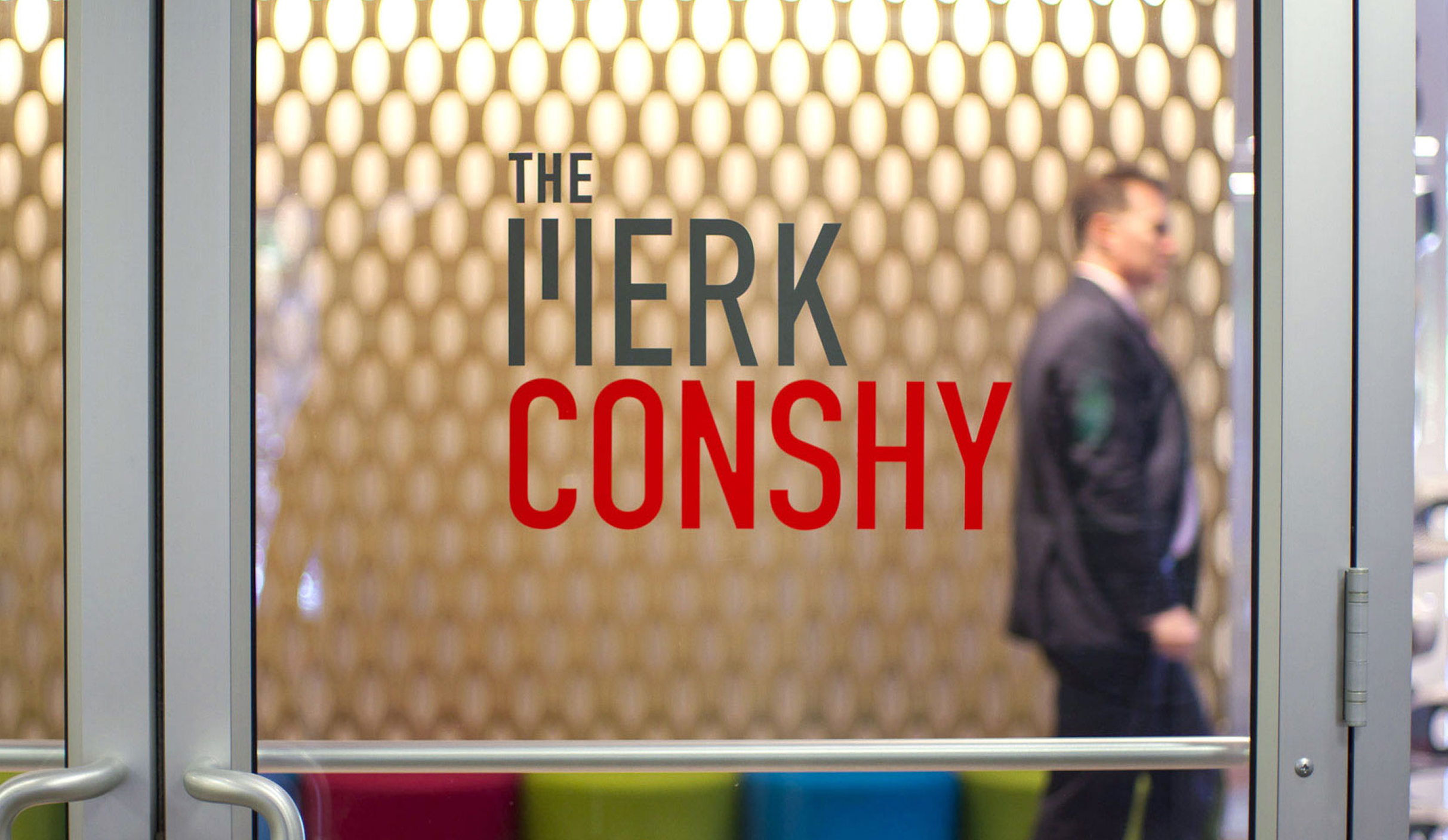 Work-Merk-Conshy A blurred man in a suit walks behind a glass door with "The MERK CONSHY" in bold letters. The background features a patterned wall and colorful seating.