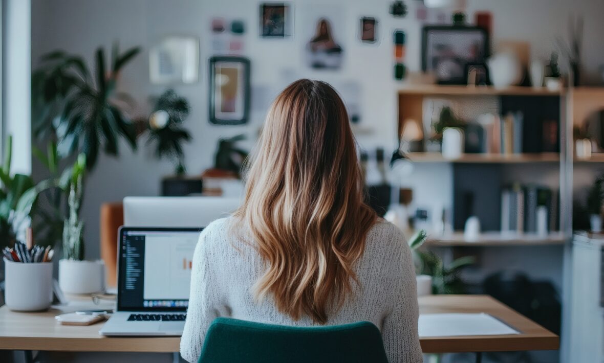 Founder working at a desk in a bright home office, viewed from behind, with a laptop open, plants, shelves, and creative materials suggesting a calm, organized workspace.