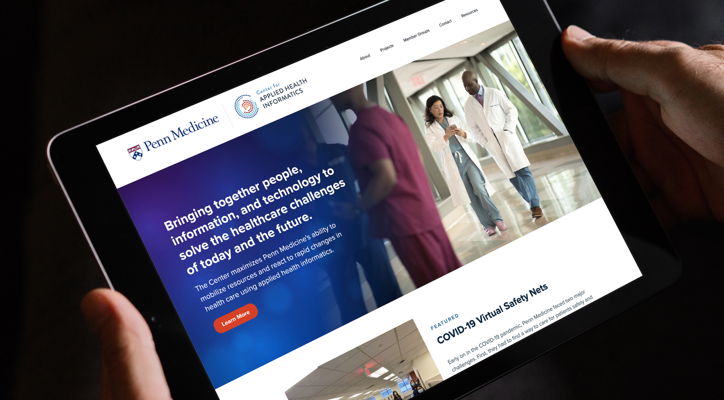 CAHI-Tablet2 Two hands hold a tablet displaying the CAHI website. The screen shows a white-coat clinician walking with a nurse. Text mentions healthcare solutions.