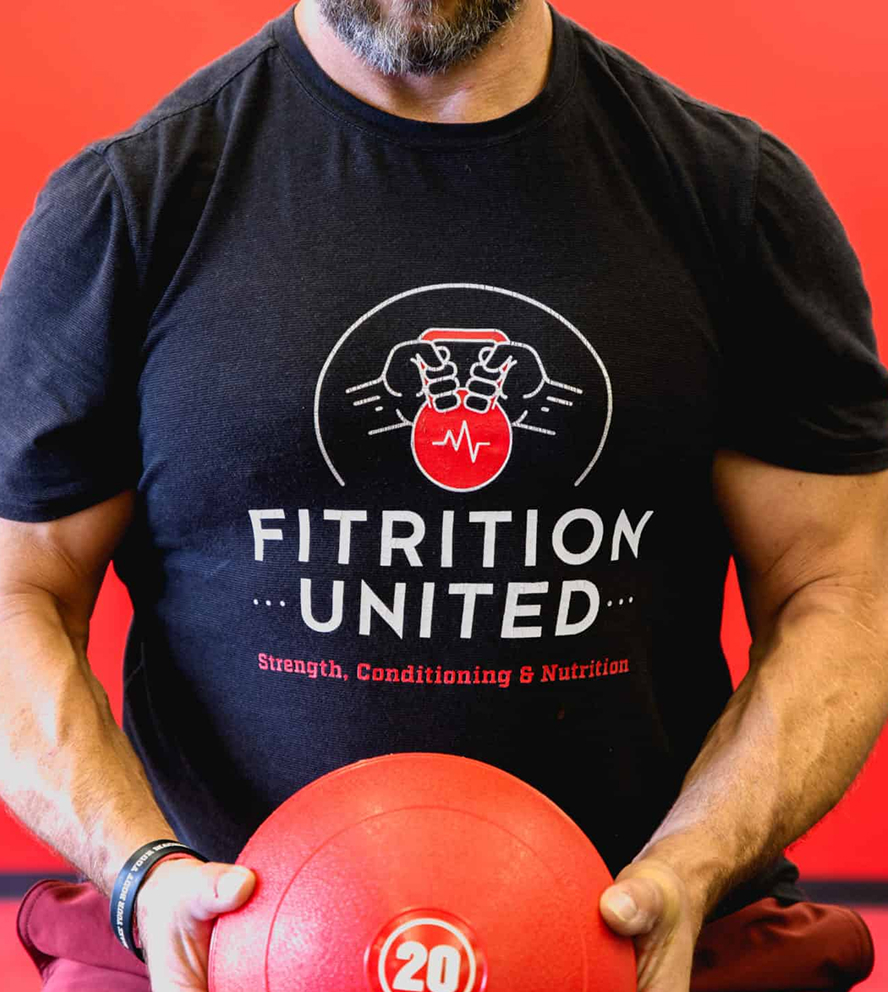 FitU-Shirt Man in a black "Fitrition United" t-shirt holds a red medicine ball. The shirt features a logo with hands gripping a kettlebell.