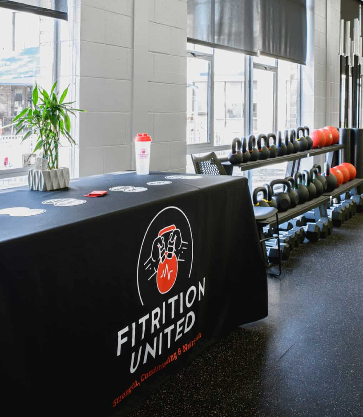 FitU-Table Bright gym room with kettlebells on racks, a table with a "Fitrition United" banner, a plant, coasters, and a shaker bottle, creating a motivational vibe.
