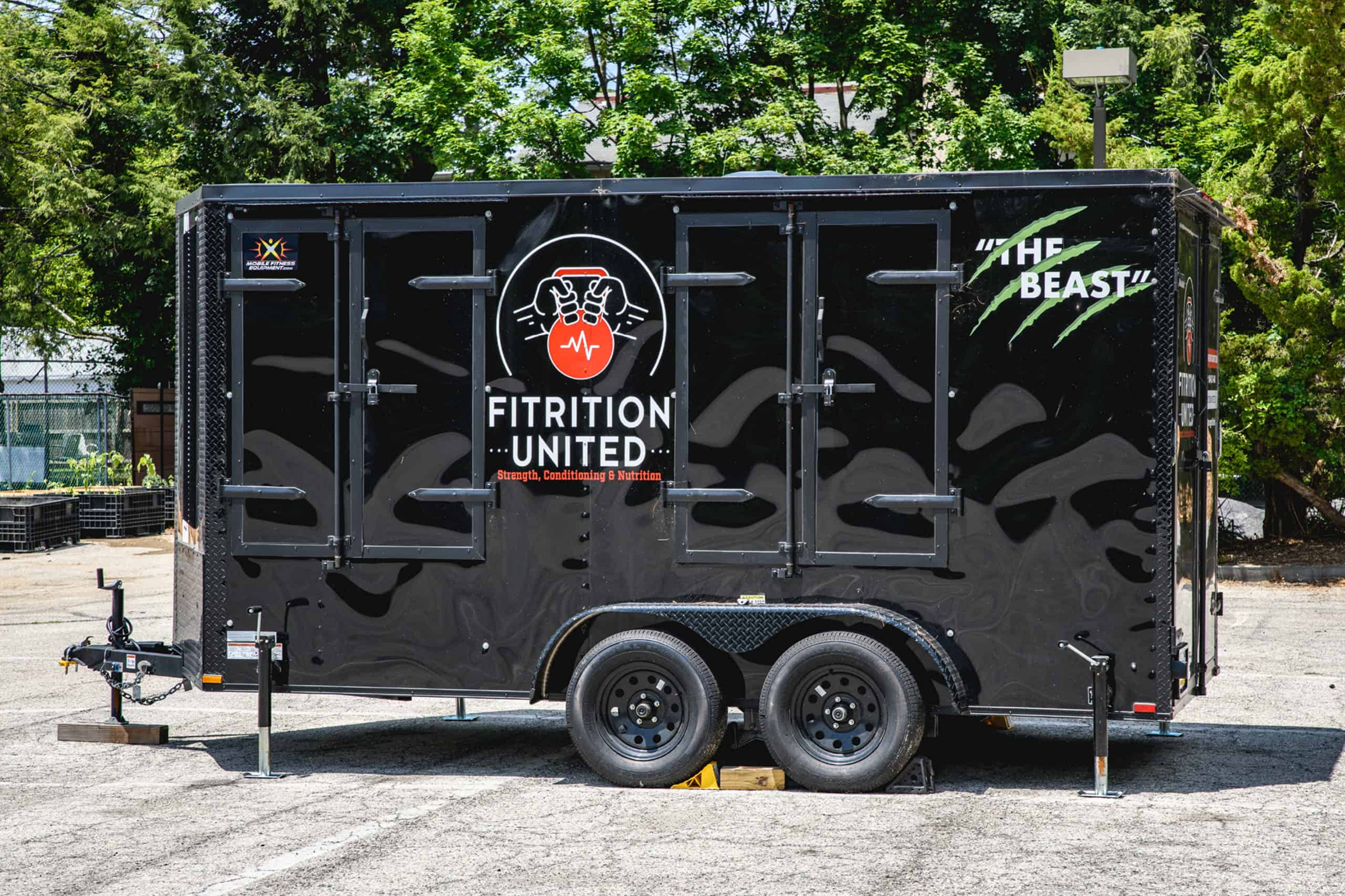 FitU-Truck Black trailer labeled "Fitrition United" with a red emblem, parked outdoors. "The Beast" is written in green with claw marks. Trees in background.