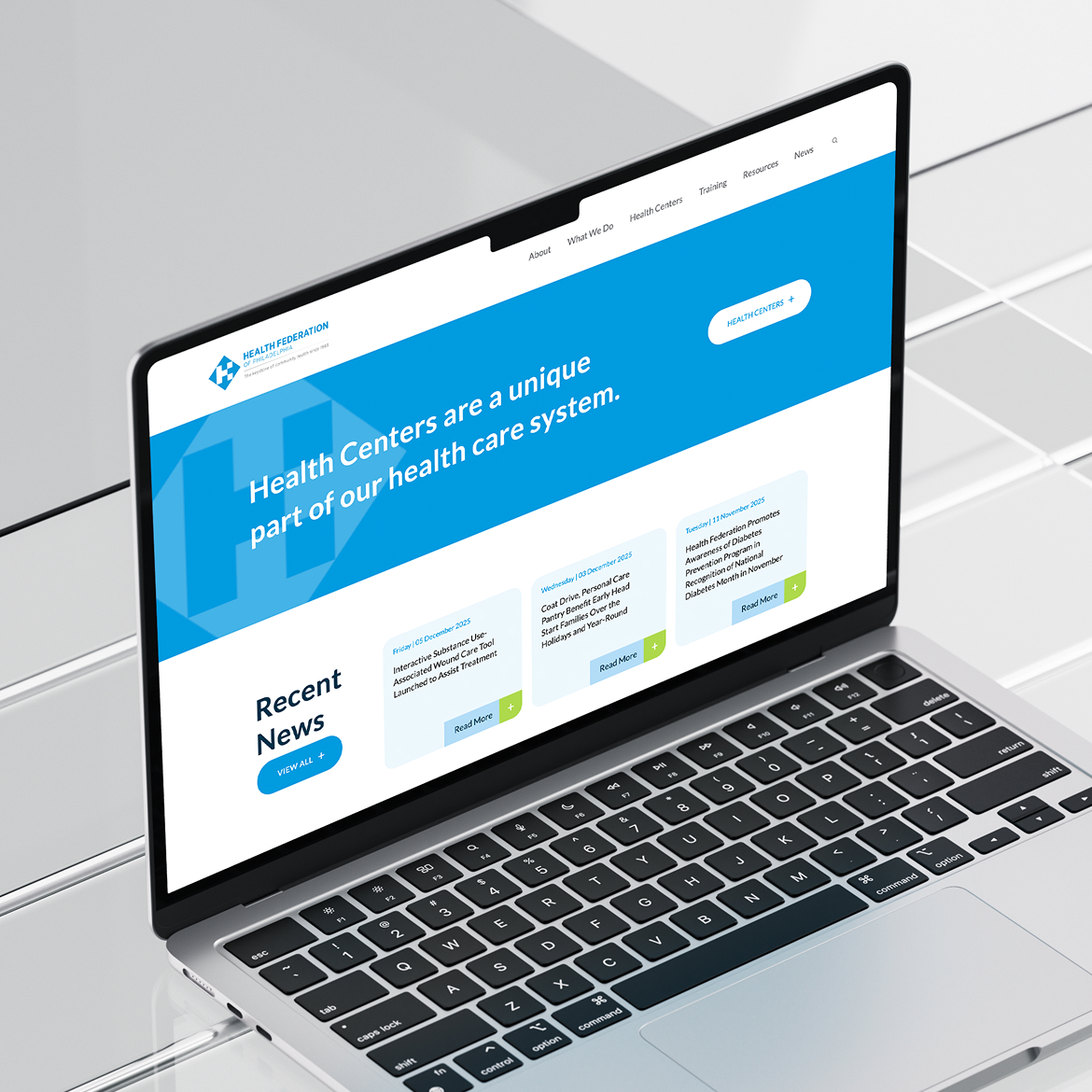 HealthFed-Website A laptop displaying the Health Federation website. The screen shows a blue banner with text, navigation menu, and recent news section. Clean, modern design.