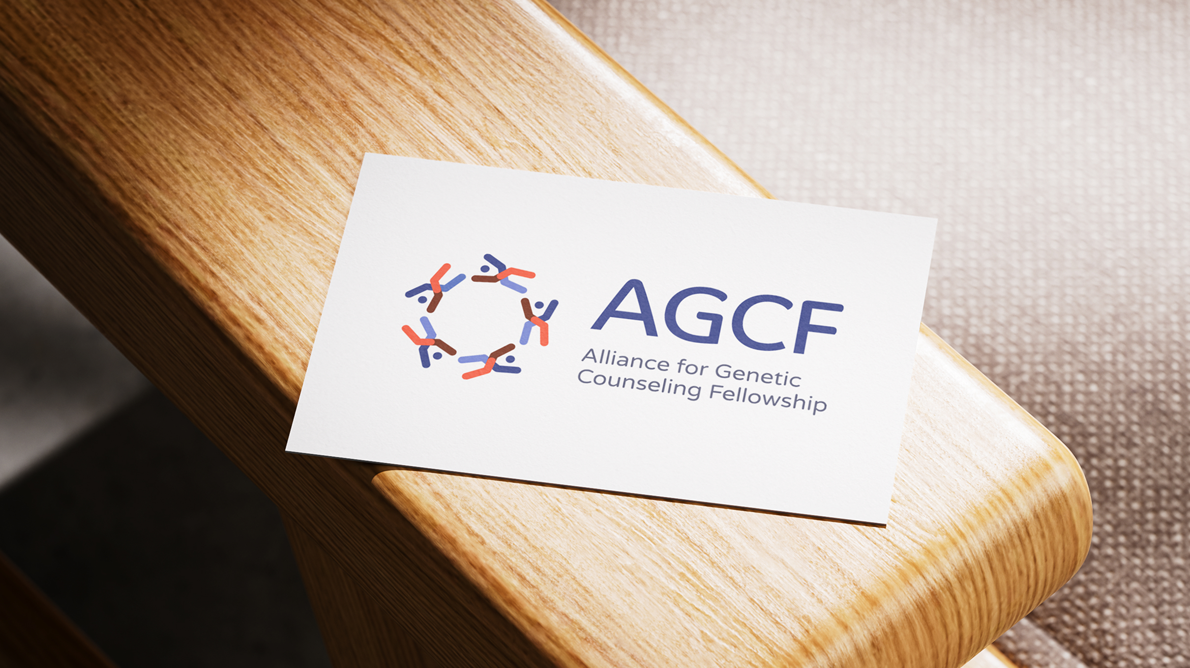 A business card on a wooden surface shows the AGCF logo. The logo features stylized figures in the shape of chromosomes, in a circle. To the right text reads "AGCF" in bold blue letters with "Alliance for Genetic Counseling Fellowship" underneath.
