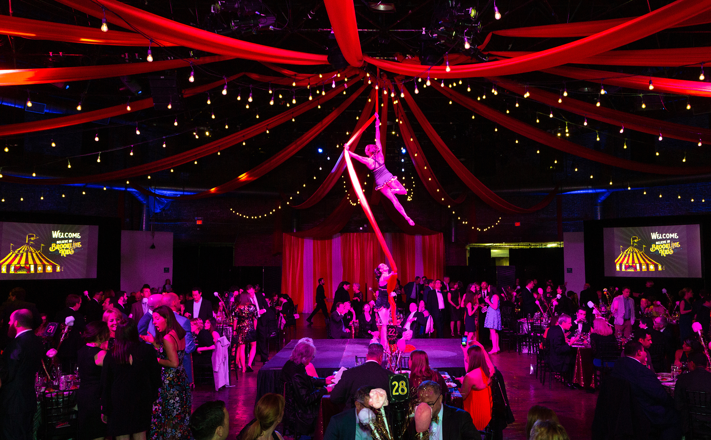 Aerial performer on red silks above a lively gala, with vibrant string lights and draped fabric overhead. Guests in formal attire socialize below.