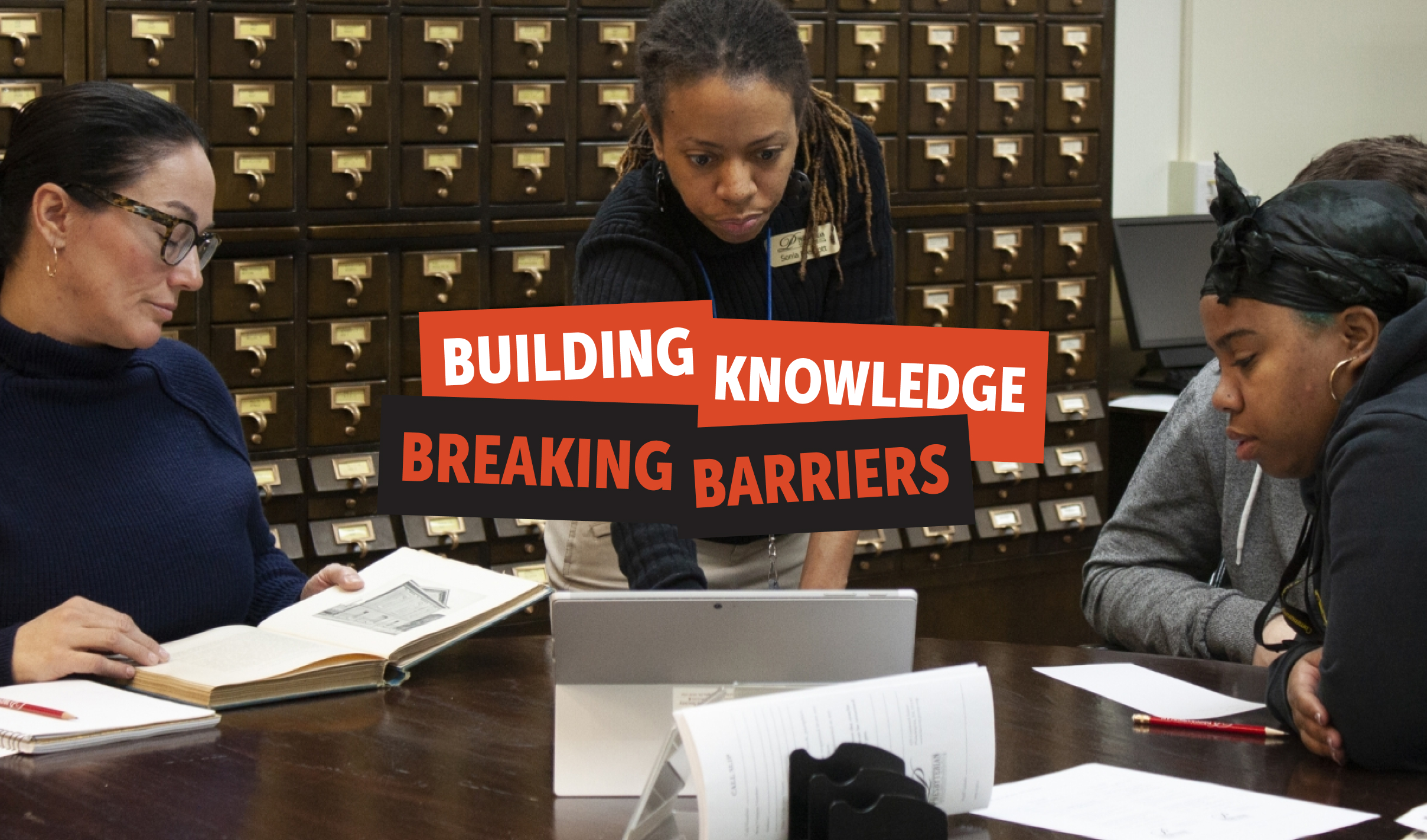BKBB-Hero A diverse group of four people in a library setting focuses intently on a tablet and open books. Red and black rectangles sit on top of the image, containing the words "Building Knowledge, Breaking Barriers."
