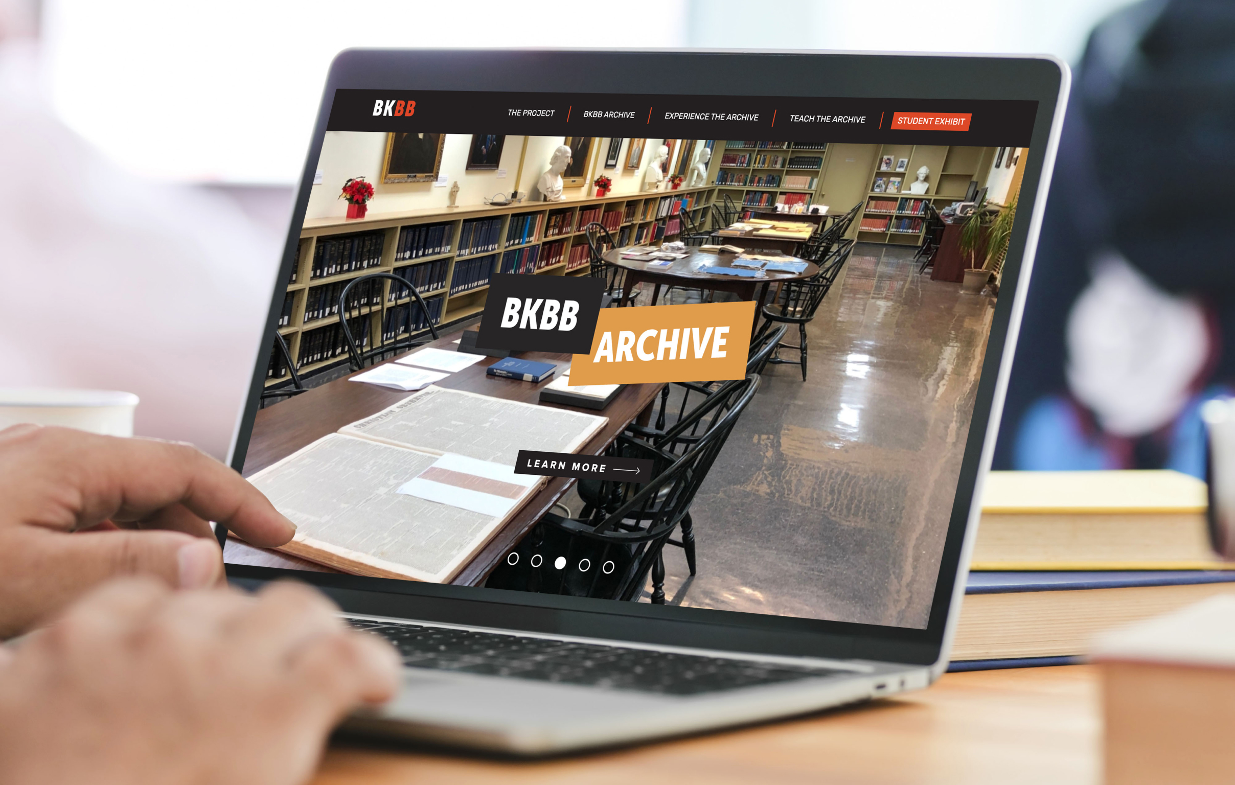 BKBB-Homepage Laptop screen displaying the BKBB archive website, featuring an image of a library interior with tables, chairs, and bookshelves. Hands are typing, suggesting research.