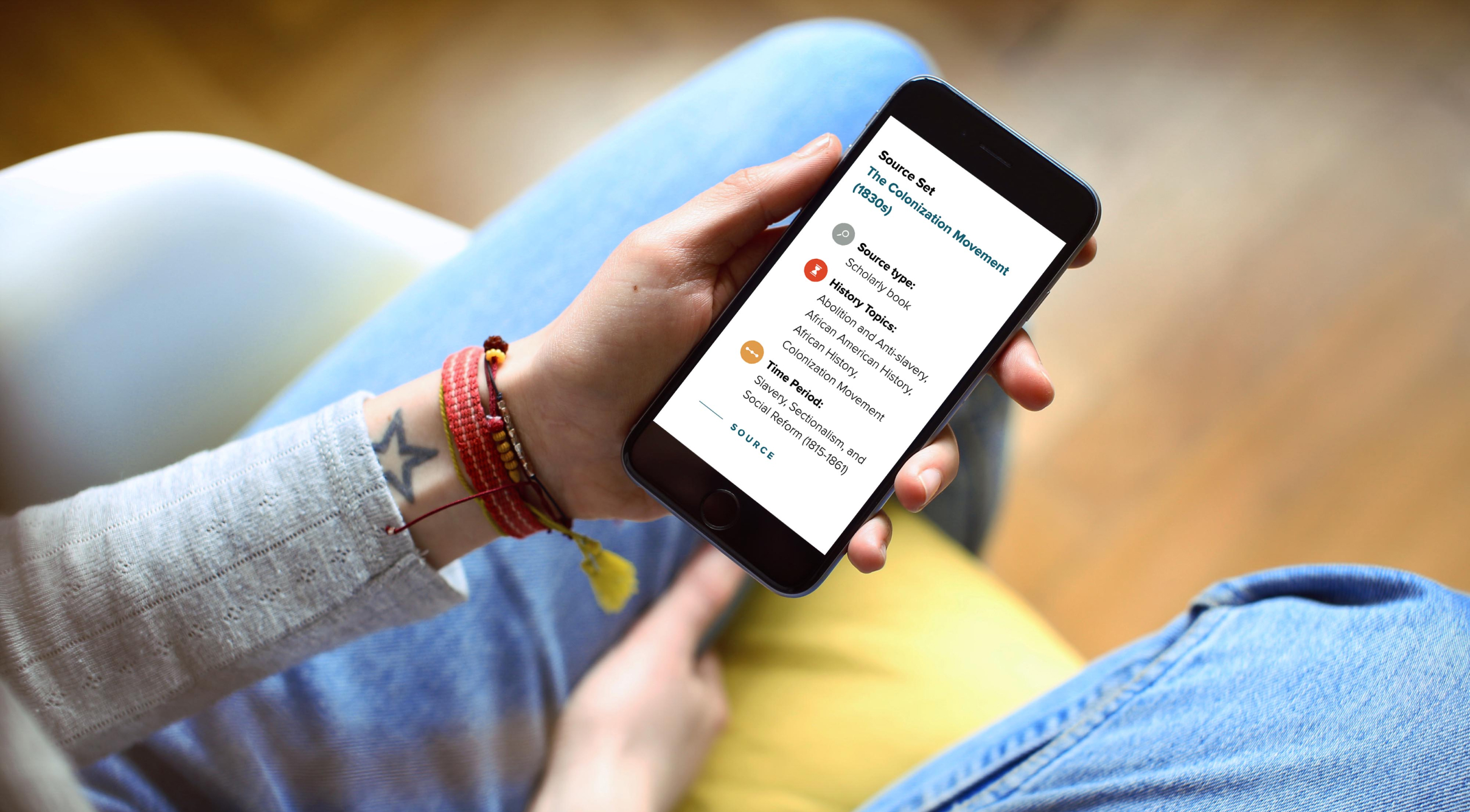 BKBB-Phone A person sits holding a smartphone in one hand. The phone screen displays an educational interface about historical sources, including topics related to colonization and history. The person wears jeans, a light-colored top, and several bracelets, with a small star tattoo visible on their wrist.