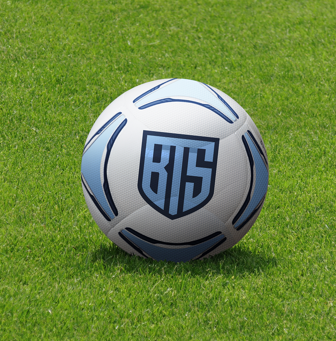 A soccer ball includes a navy blue shield logo with the letters "BTS" in bright blue. The ball sits on vibrant green grass, conveying a sense of outdoor activity and sportsmanship.