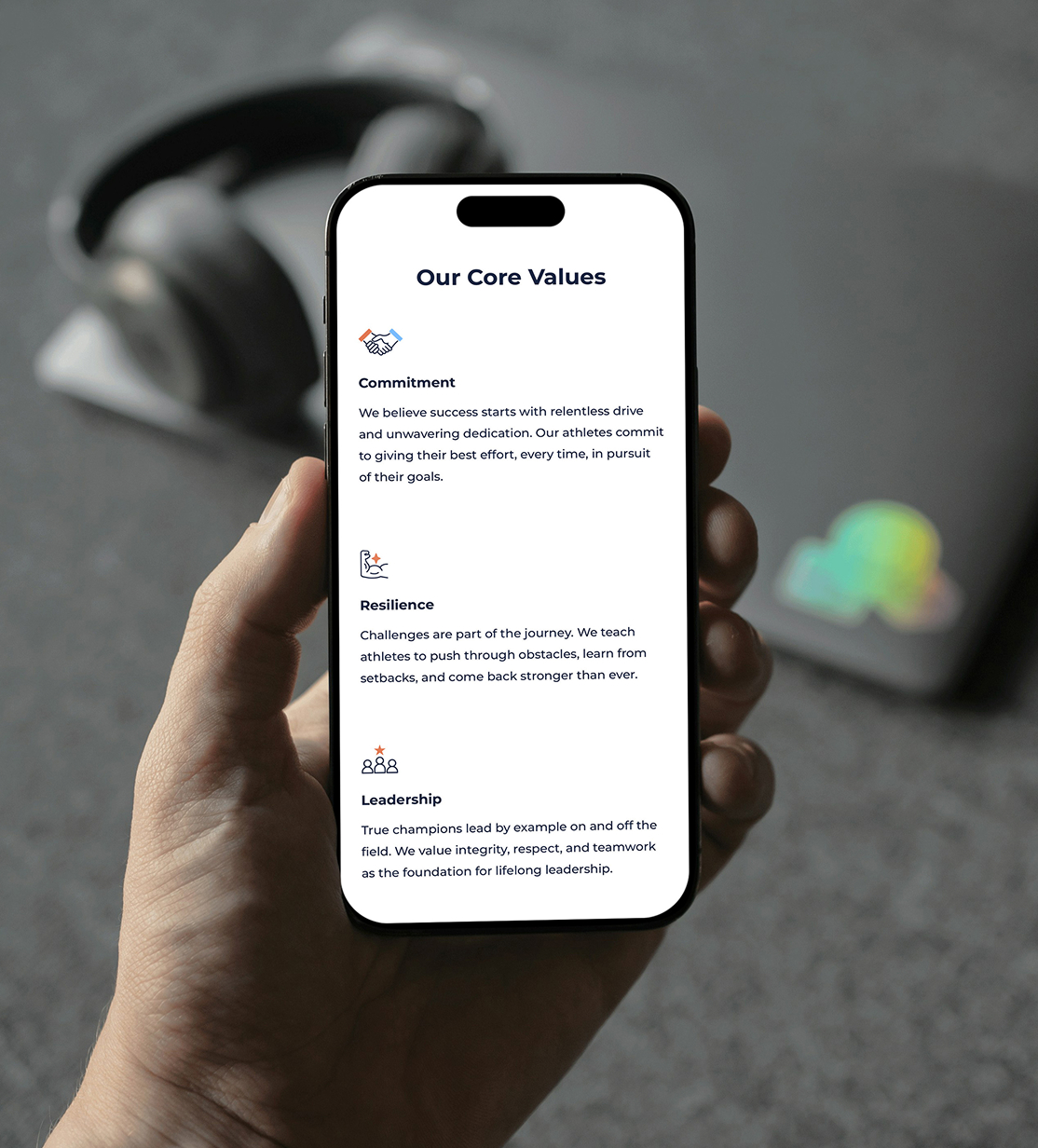 A hand holds a smartphone displaying a section from the Beyond The Season website. The title "Our Core Values" appears at the top with sections labeled "Commitment," "Resilience," and "Leadership" with accompanying icons. Blurred headphones in the background.