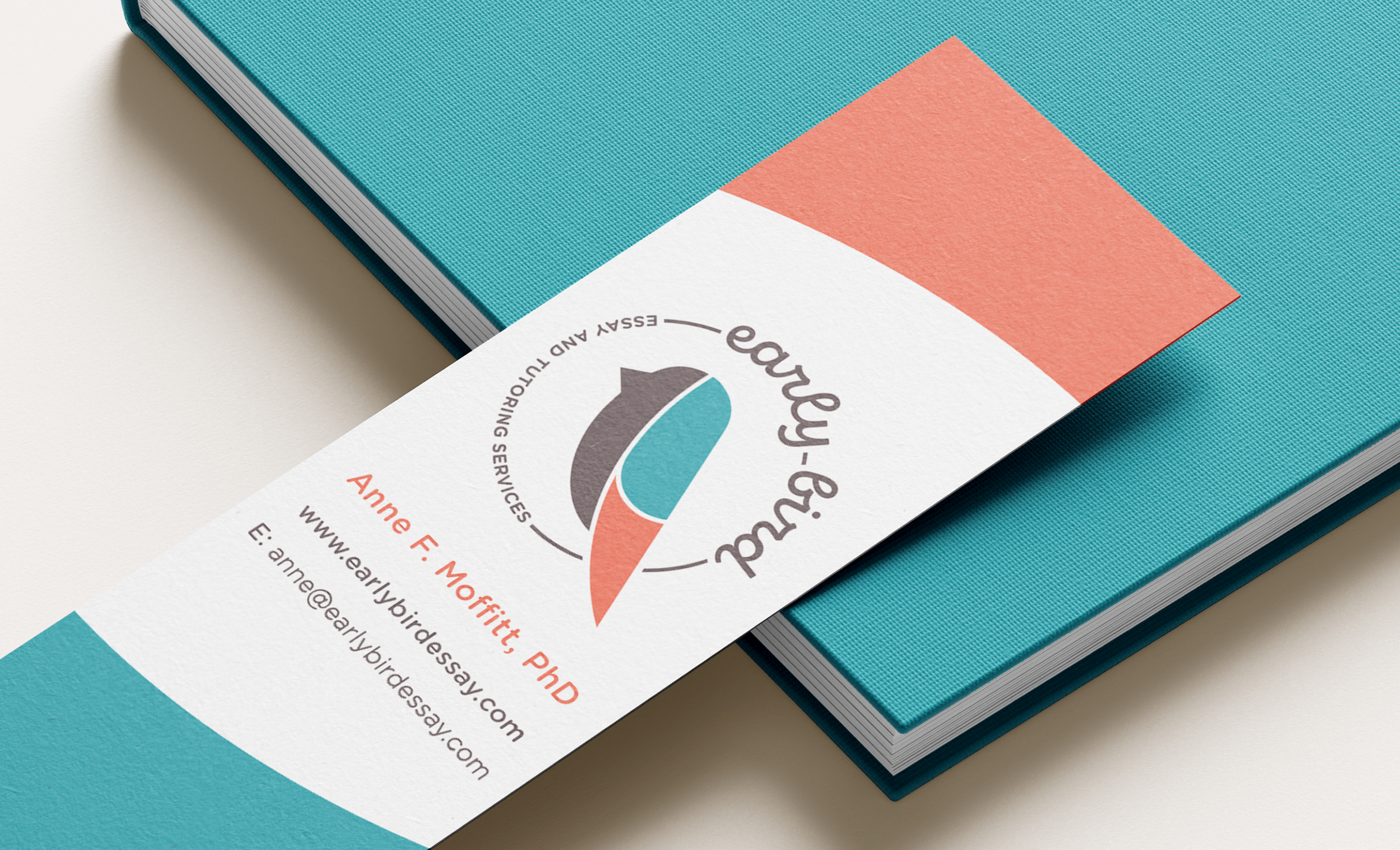 A bookmark rests on top of a teal notebook. The bookmark features color of red and blue blocks at the top and bottom, the Early Bird Tutoring logo in the center, and contact details. The logo includes a stylized bird in brown, blue, and red. In a circle around the bird text reads "early bird" in cursive, with "Essay and Tutoring Services."