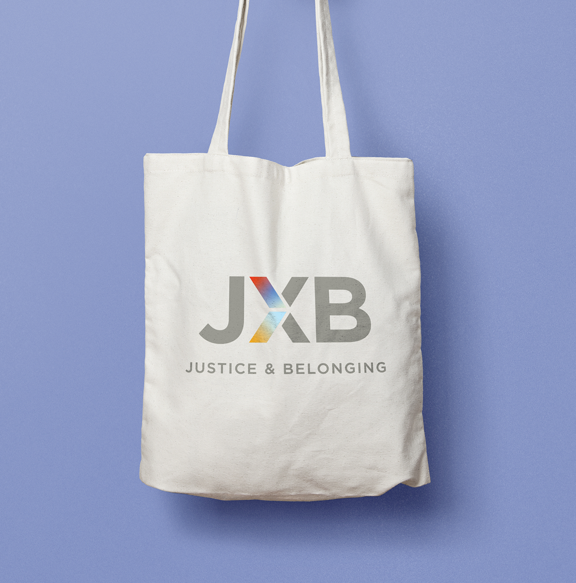 A white tote bag against a light blue background displays the JXB logo. The logo has the letters "JXB" with a colorful "X" in red, blue, and yellow hues. Below, the text reads "Justice & Belonging."