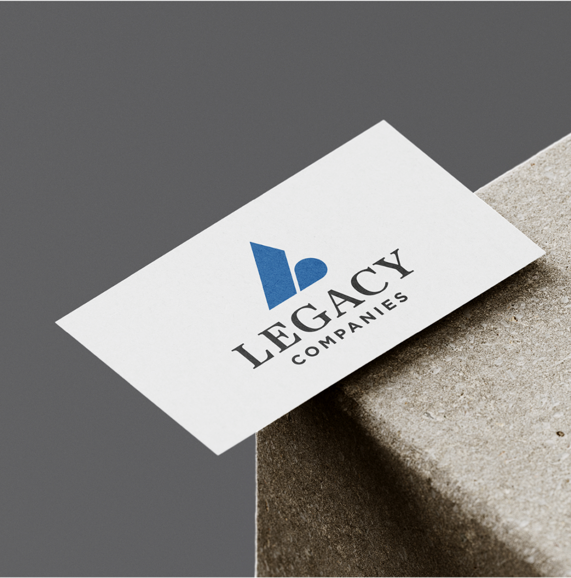 Legacy-BusinessCard White business card with a blue abstract logo and "Legacy Companies" in bold black text, resting on a textured gray surface. Elegant and professional tone.