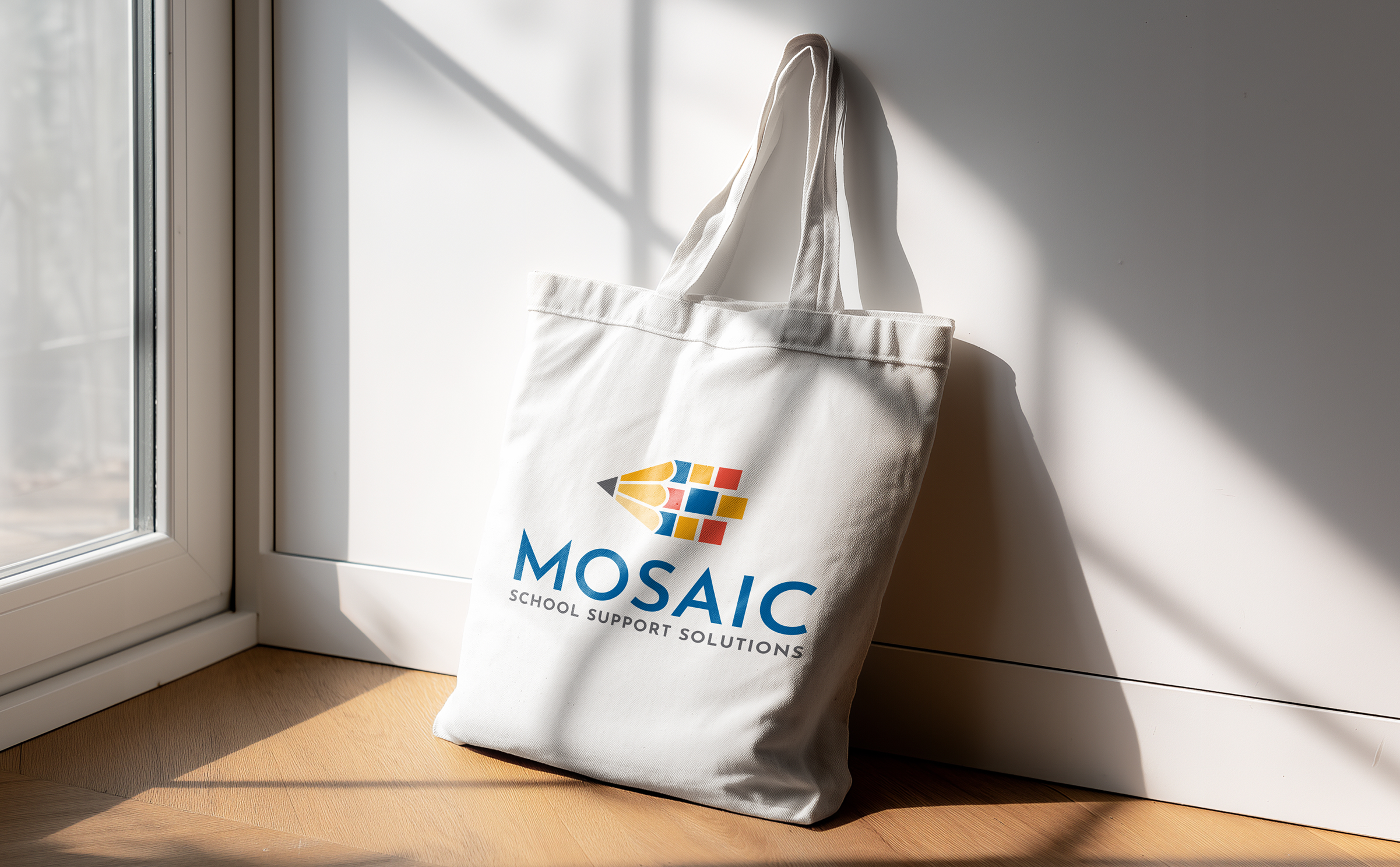 A white tote bag with the "Mosaic School Support Solutions" logo, featuring a multicolored pencil design, rests against a sunlit wall on a wooden floor.
