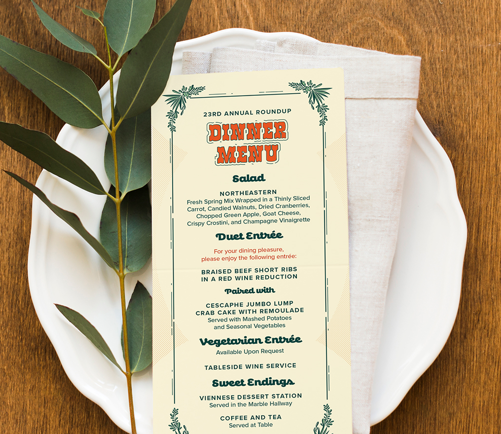 Roundup-Menu Dinner menu on off-white card with orange decorative text, placed on a white plate atop a beige napkin. Green leaves placed next to the menu add a fresh touch.