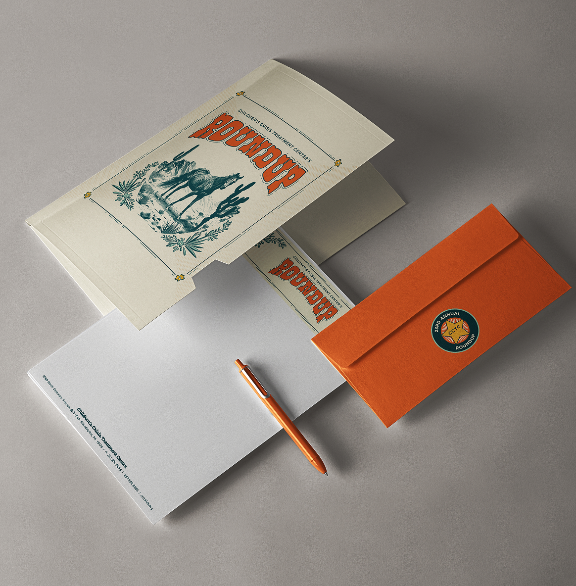 Roundup-Stationery A stationery set for CCTC's Roundup event with a western theme includes a beige folder featuring a horse illustration, an orange envelope, and a matching pen on a gray background.