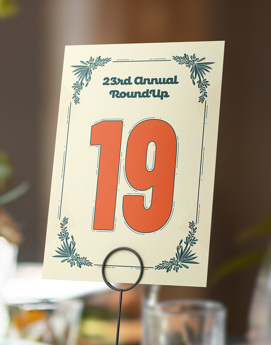Roundup-TableNumbers Table card with '23rd Annual Roundup' text and large orange '19' at center. Decorative foliage accents border the card on cream background.