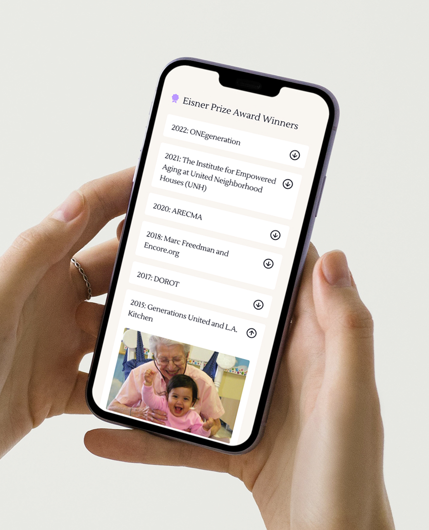 A person holds a smartphone displaying Eisner Prize Award Winners list. An image below shows a joyful elderly person hugging a smiling child.