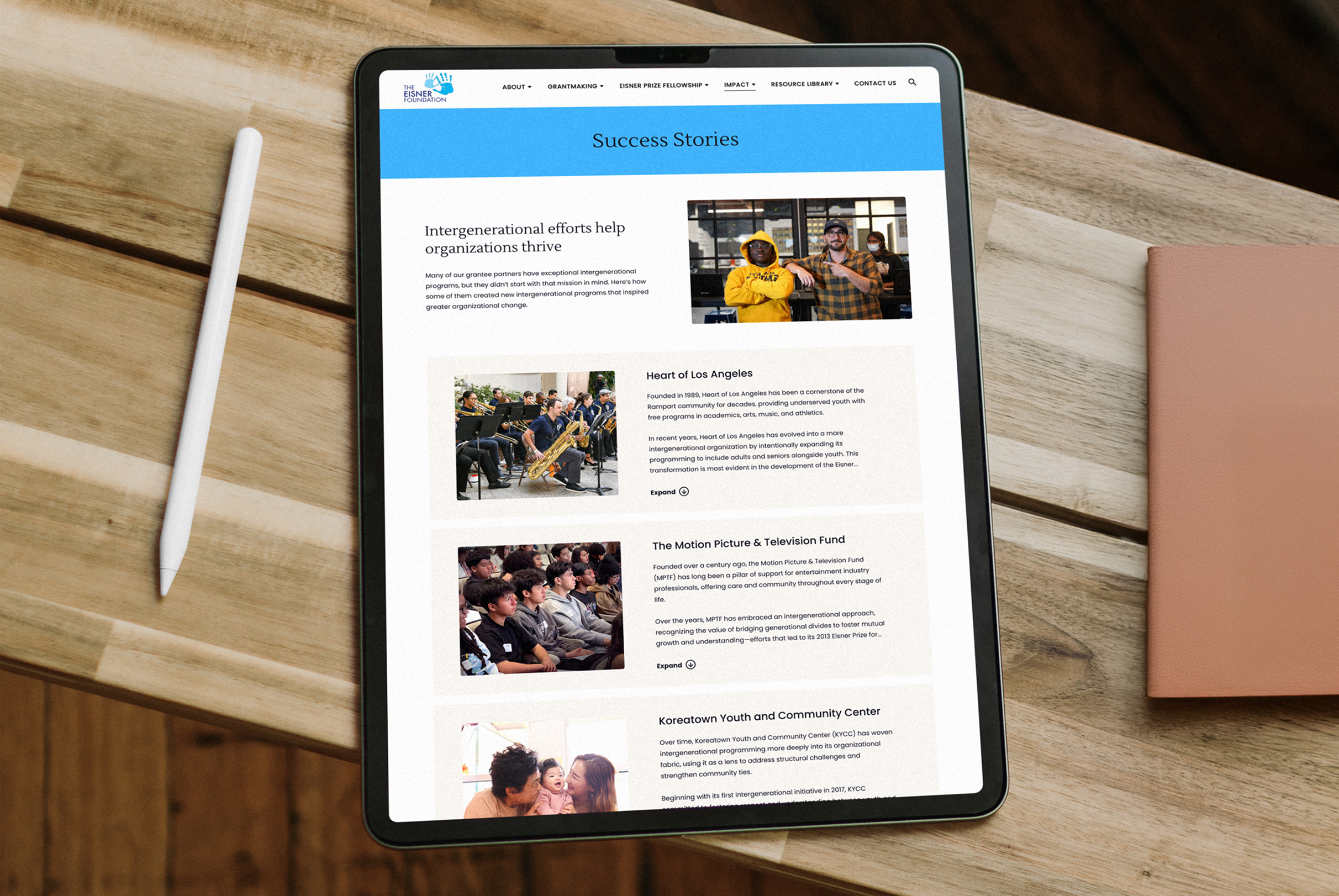 A tablet displaying the "Success Stories" page from the Eisner Foundation website with sections on intergenerational efforts. The page showcases photos and text about various community programs. A stylus and notebook lie nearby.