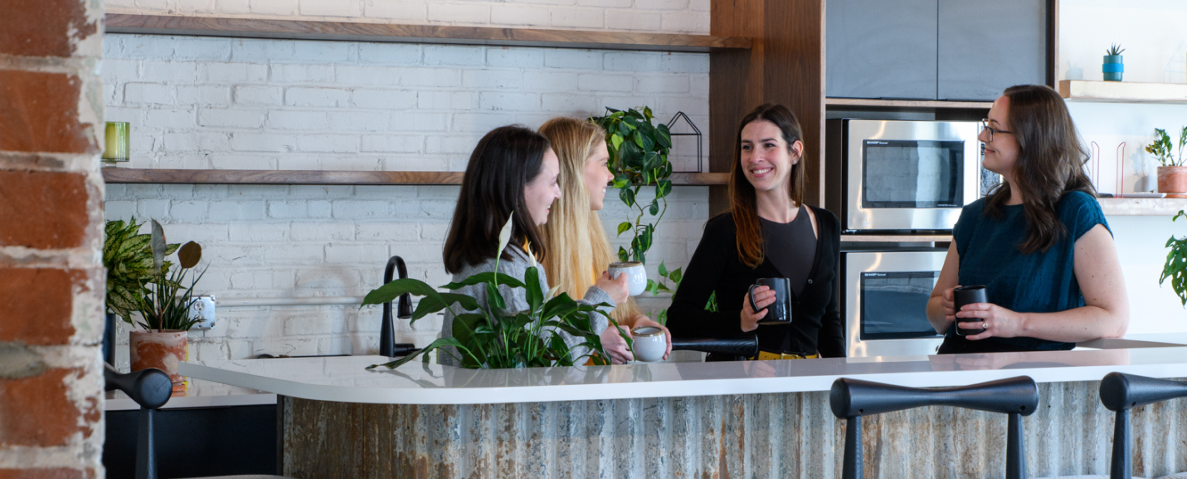 Frame 6 Four women chat and smile around a kitchen island, surrounded by plants, brick walls, modern appliances, and bar stools.